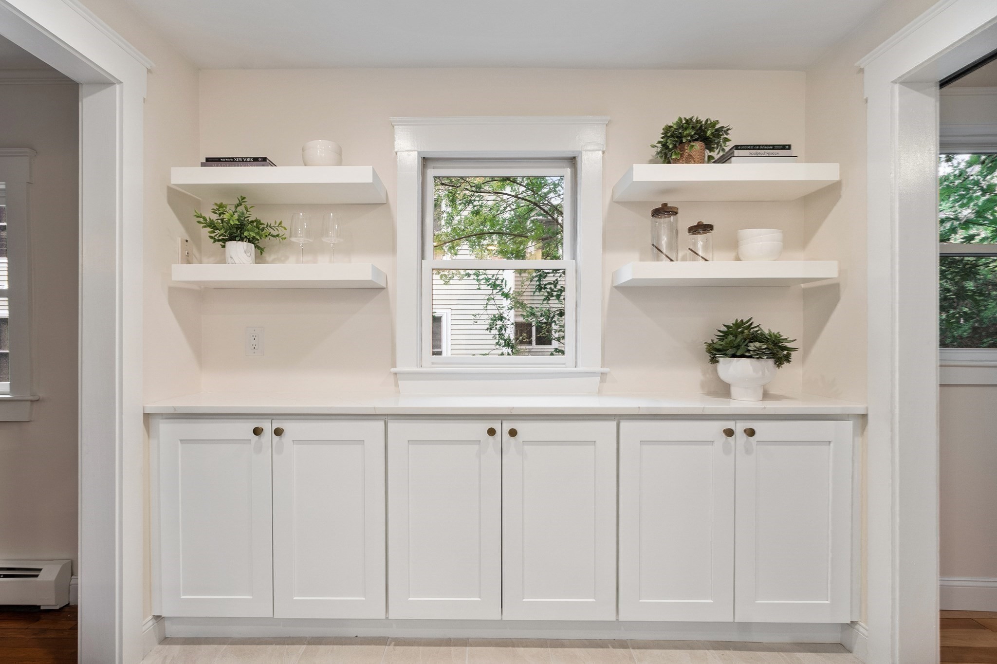 White kitchen cabinetry and floating shelves frame a window overlooking leafy trees.
