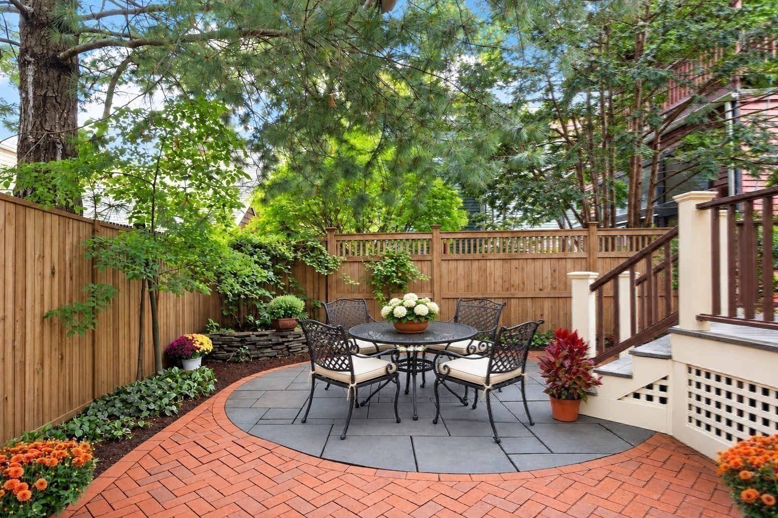 Backyard patio with a round dining set, brick paving, flower beds, and leafy trees.