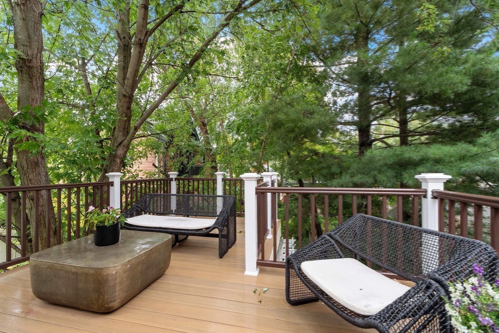Tree-shaded wooden deck with woven benches, white cushions, and a low stone coffee table.