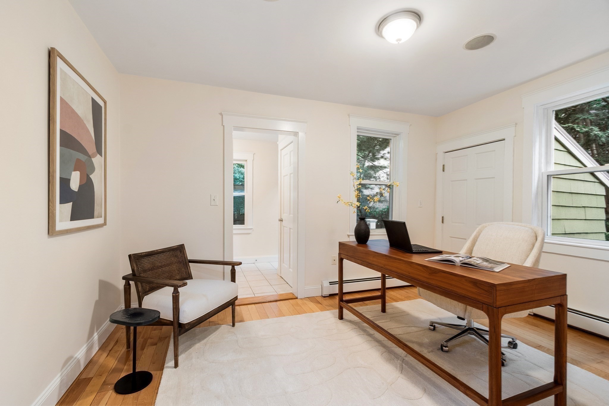 Furnished home office with a wooden desk, laptop, armchairs, and large windows.