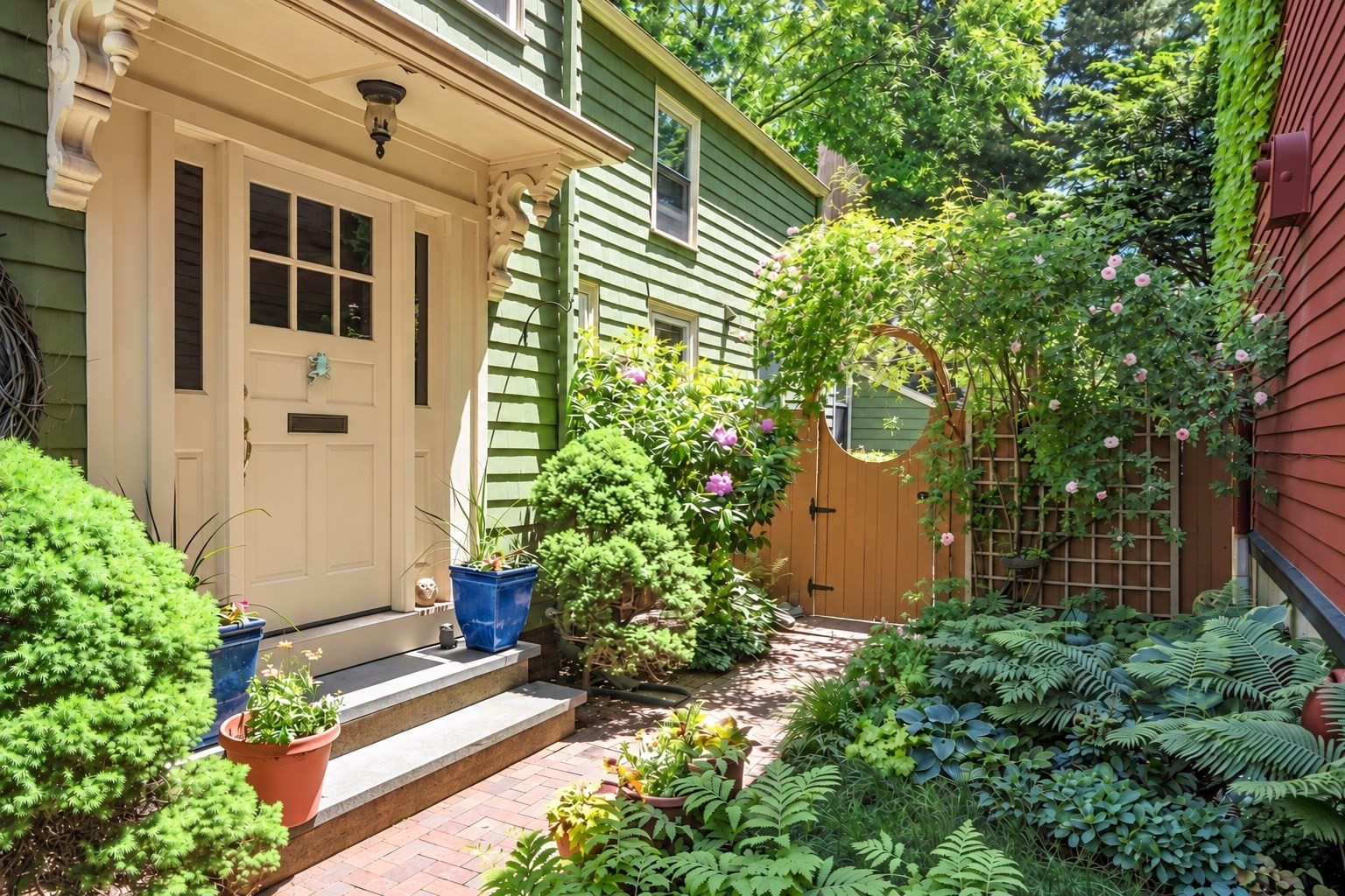 Green-shingled home entrance with a cream door, brick path, flowering garden, and wooden gate.