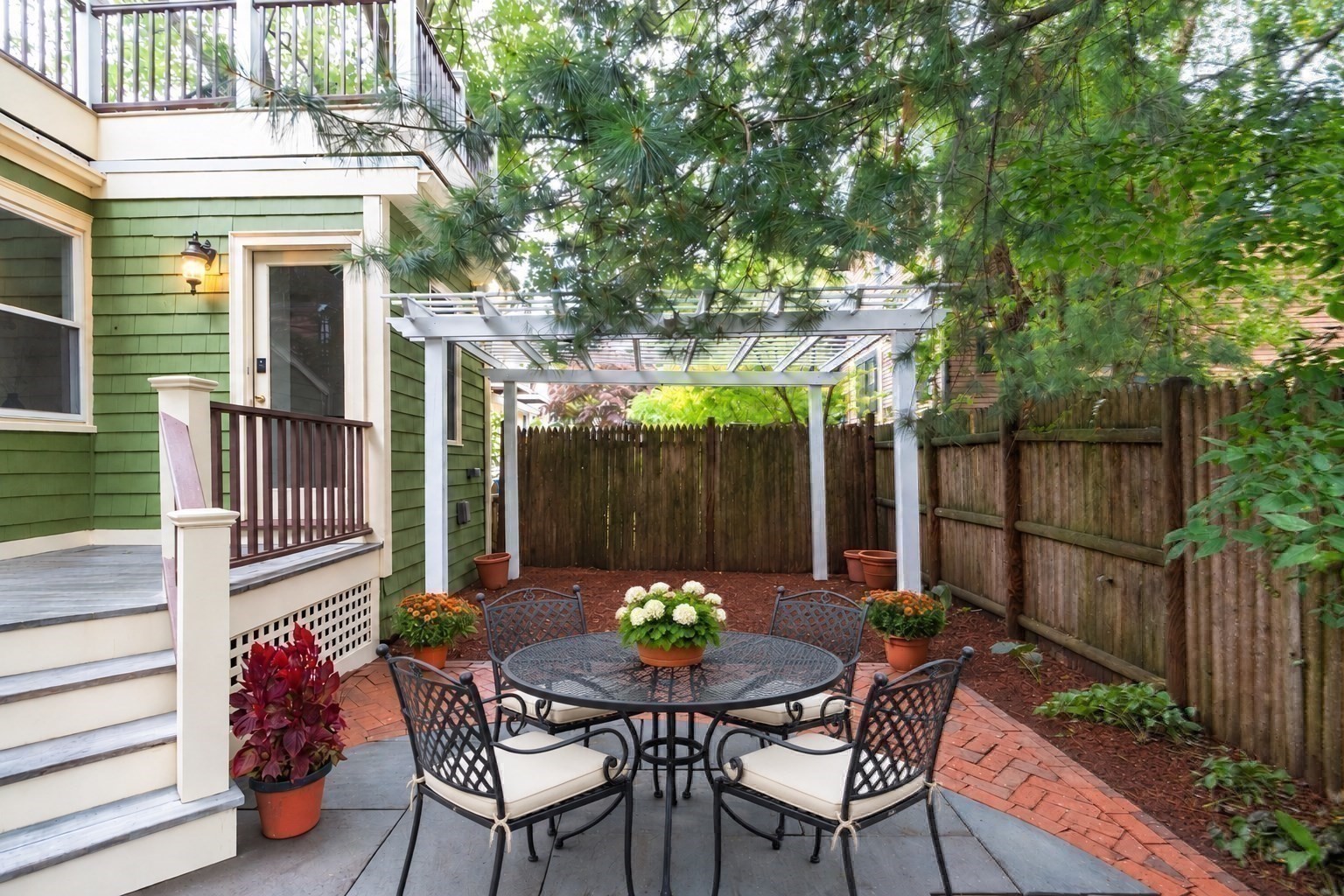 Backyard patio with a round metal dining set beneath a white pergola, surrounded by leafy trees and a wooden fence.