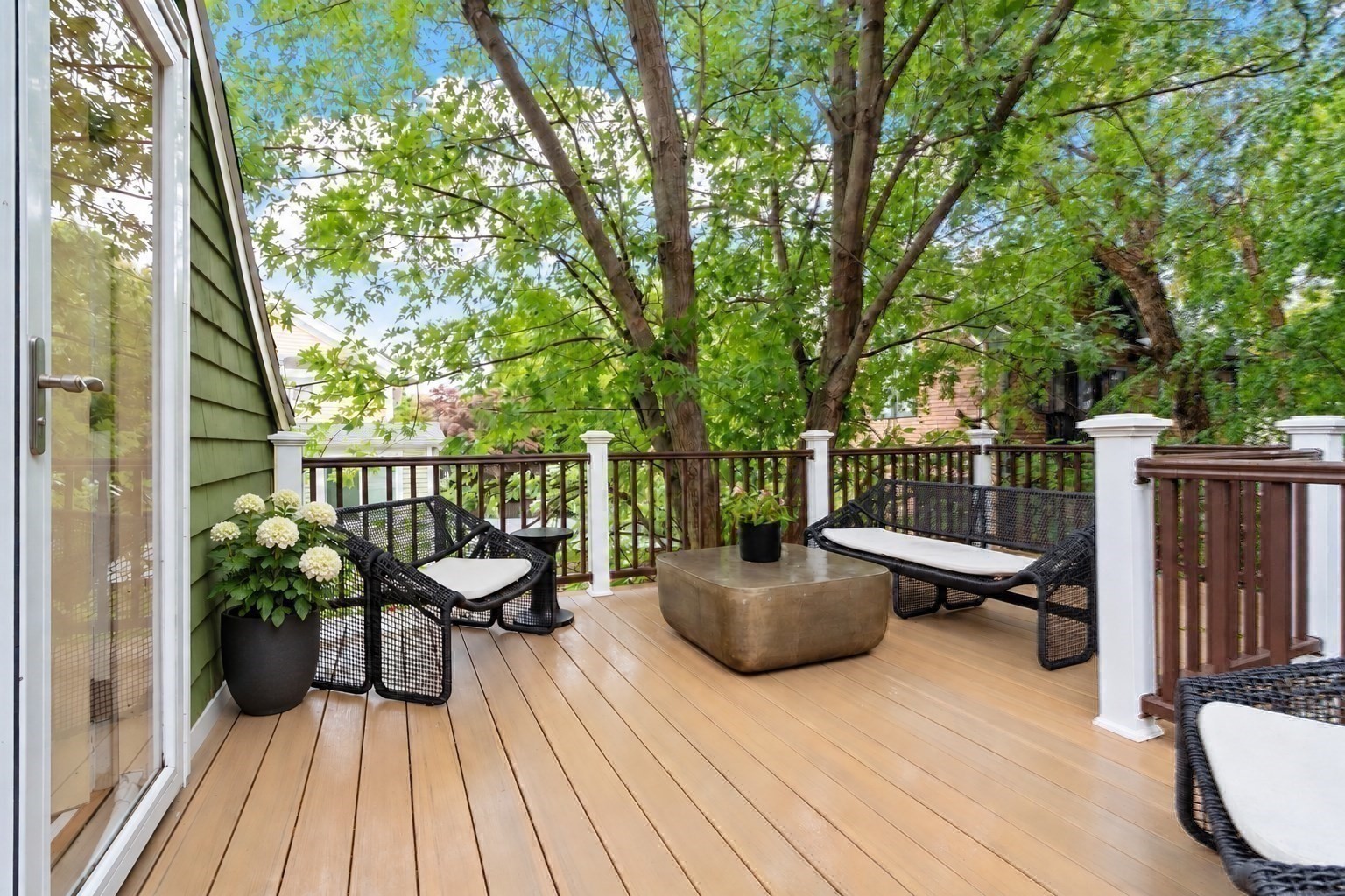 Wooden deck with woven lounge chairs and a low table beneath leafy trees.