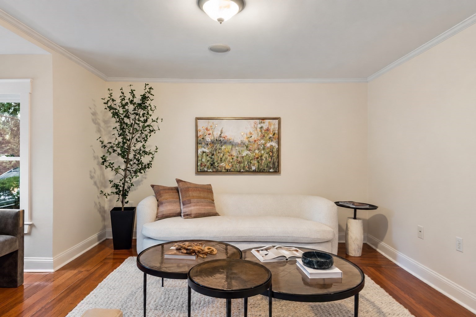 Staged living room with a white curved sofa, nesting coffee tables, and a tall potted tree.