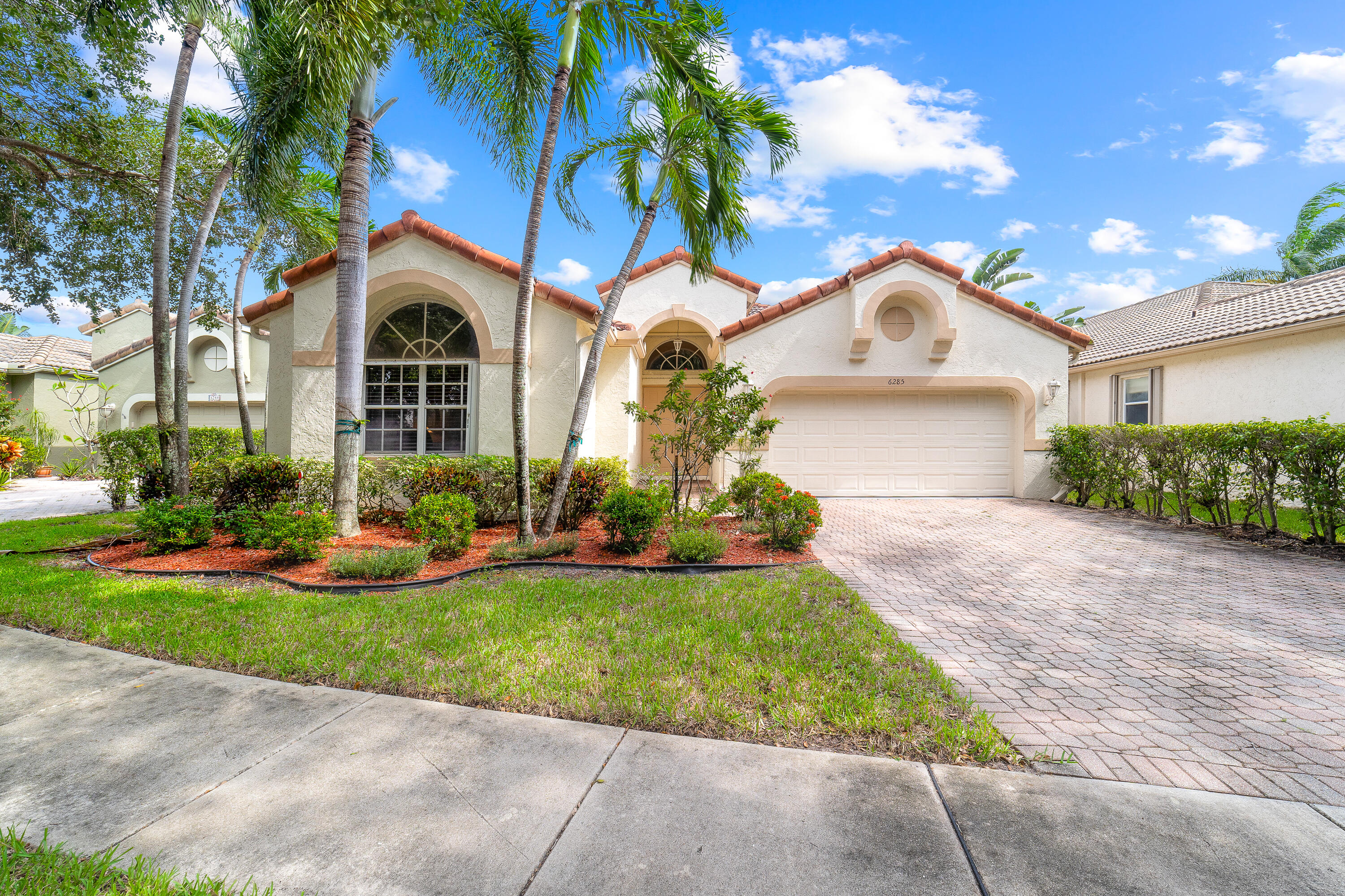 Cream stucco home with terracotta roof, arched windows, palm trees, and a wide paver driveway.