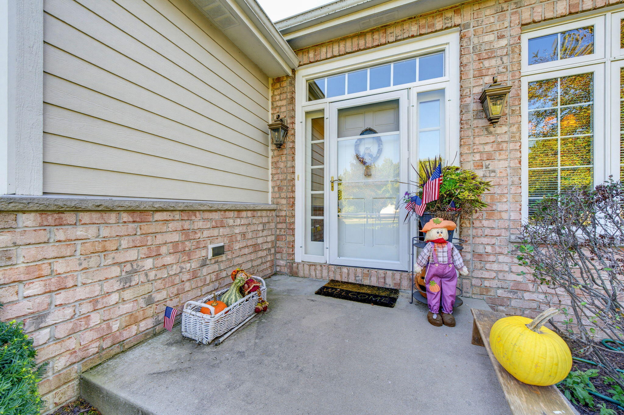 Covered brick front entry with a glass door, scarecrow, pumpkins, and fall decorations.