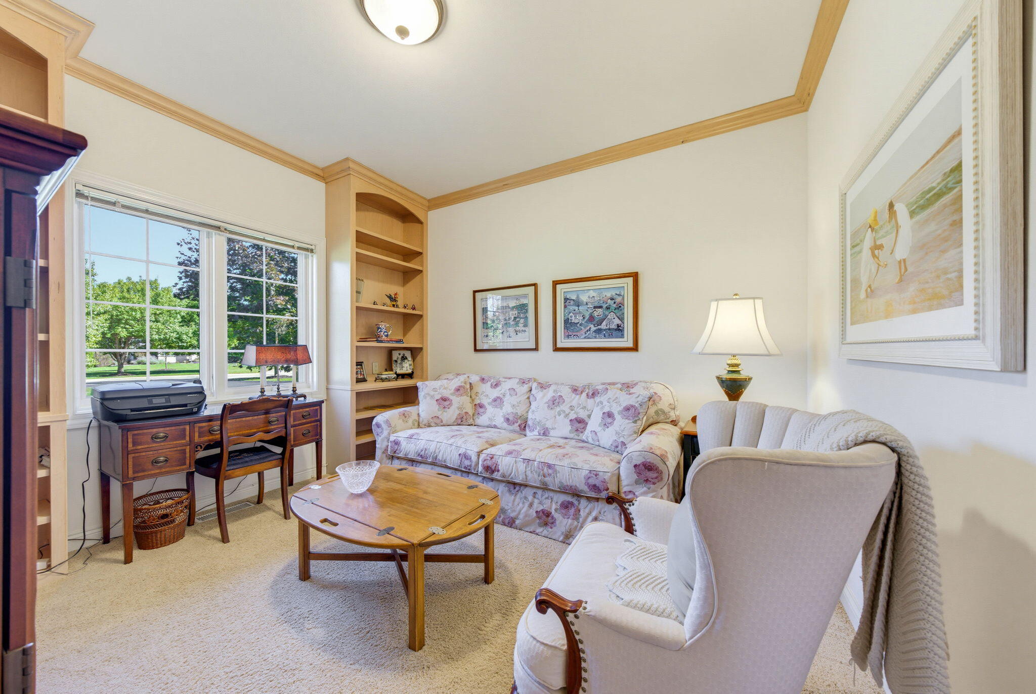 Furnished sitting room with built-in shelves, a floral sofa, and windows overlooking a green lawn.