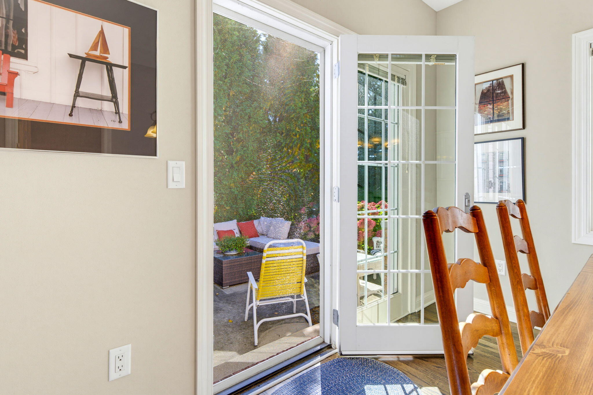 Dining area with wooden chairs beside an open French door overlooking a leafy patio.