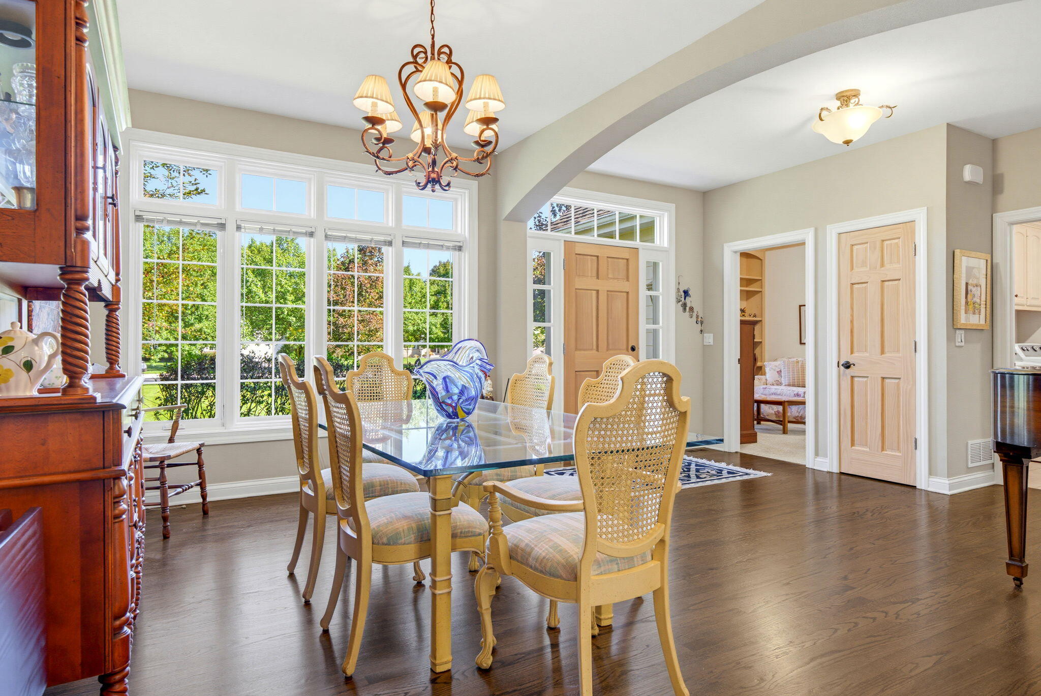 Dining room with a glass-top table, cane-back chairs, and large windows overlooking greenery.