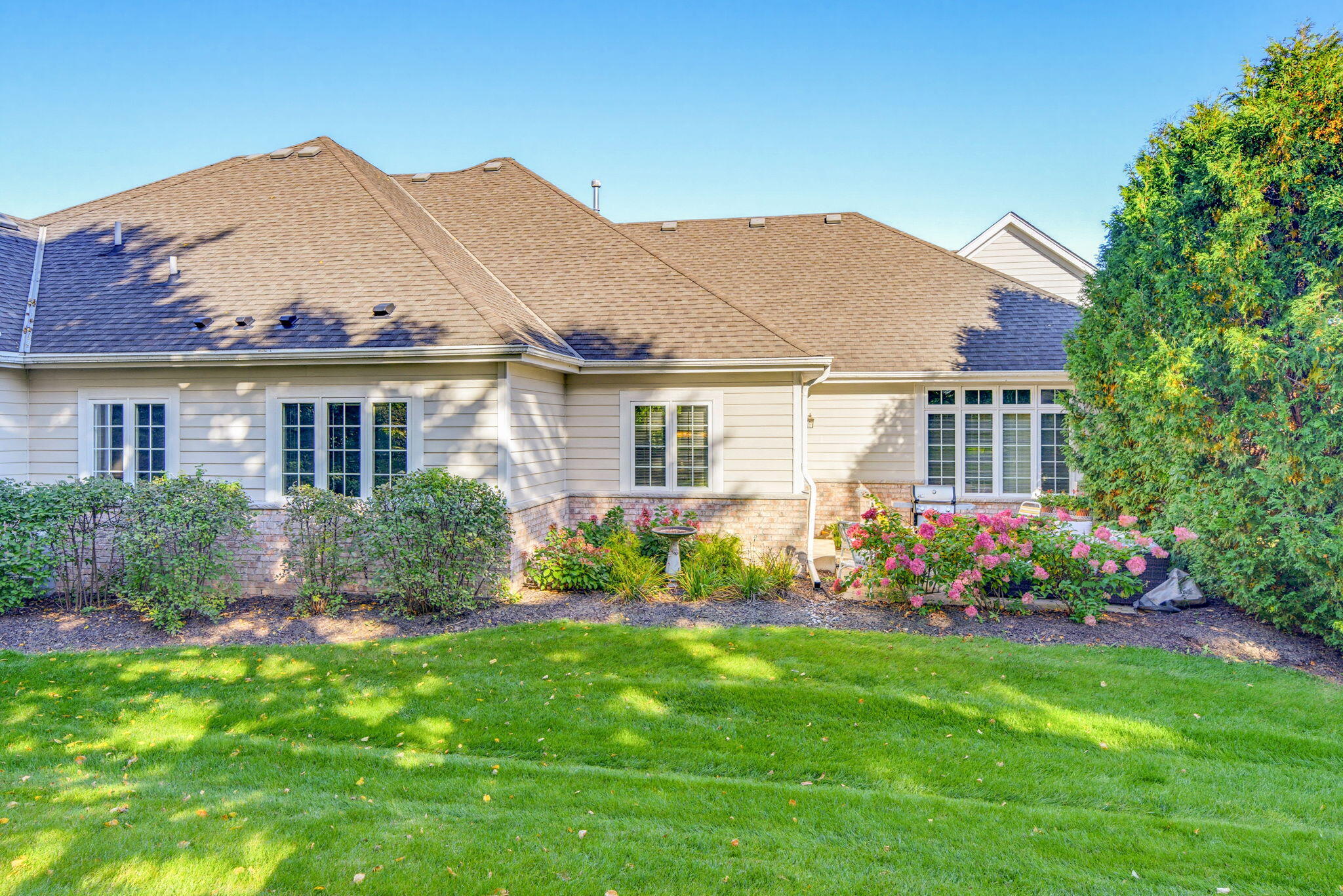 Rear of a light-sided home with pink flowering shrubs, landscaped beds, and a broad green lawn.