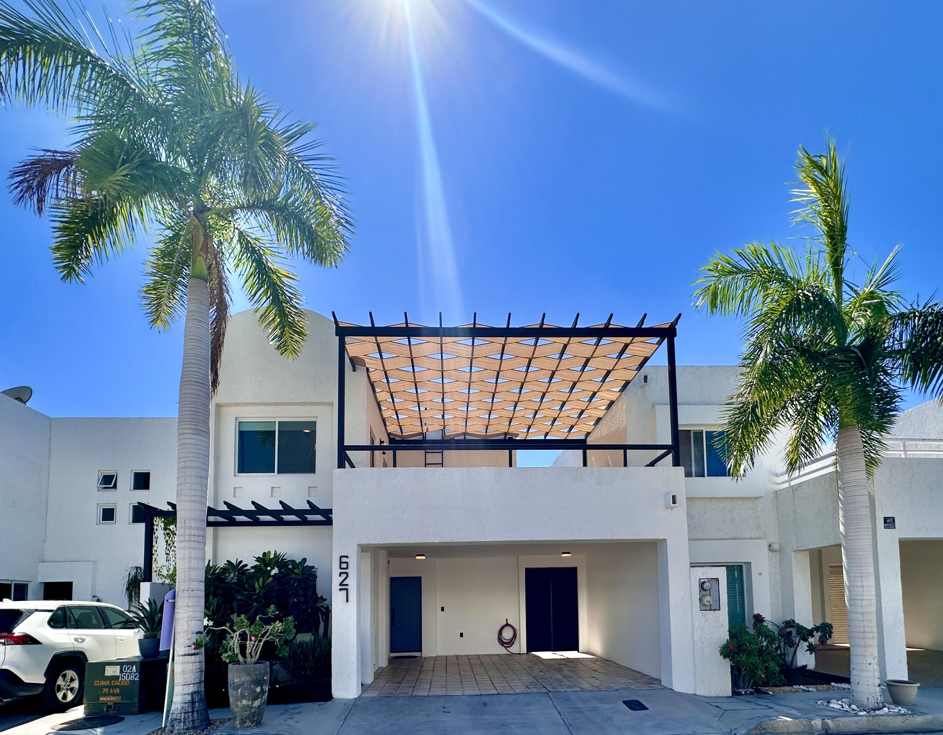 Modern white home with a geometric pergola, covered driveway, and tall palm trees.