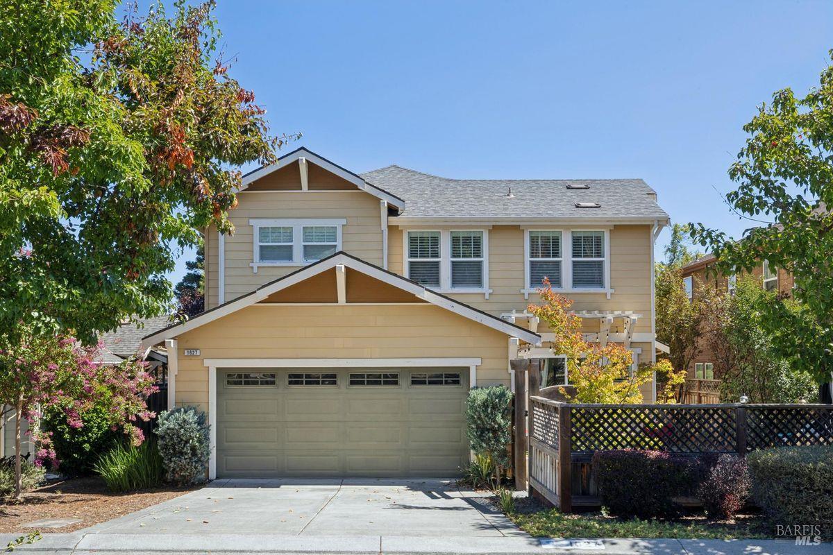 Two-story tan house with a wide double garage, landscaped front yard, and lattice fence.