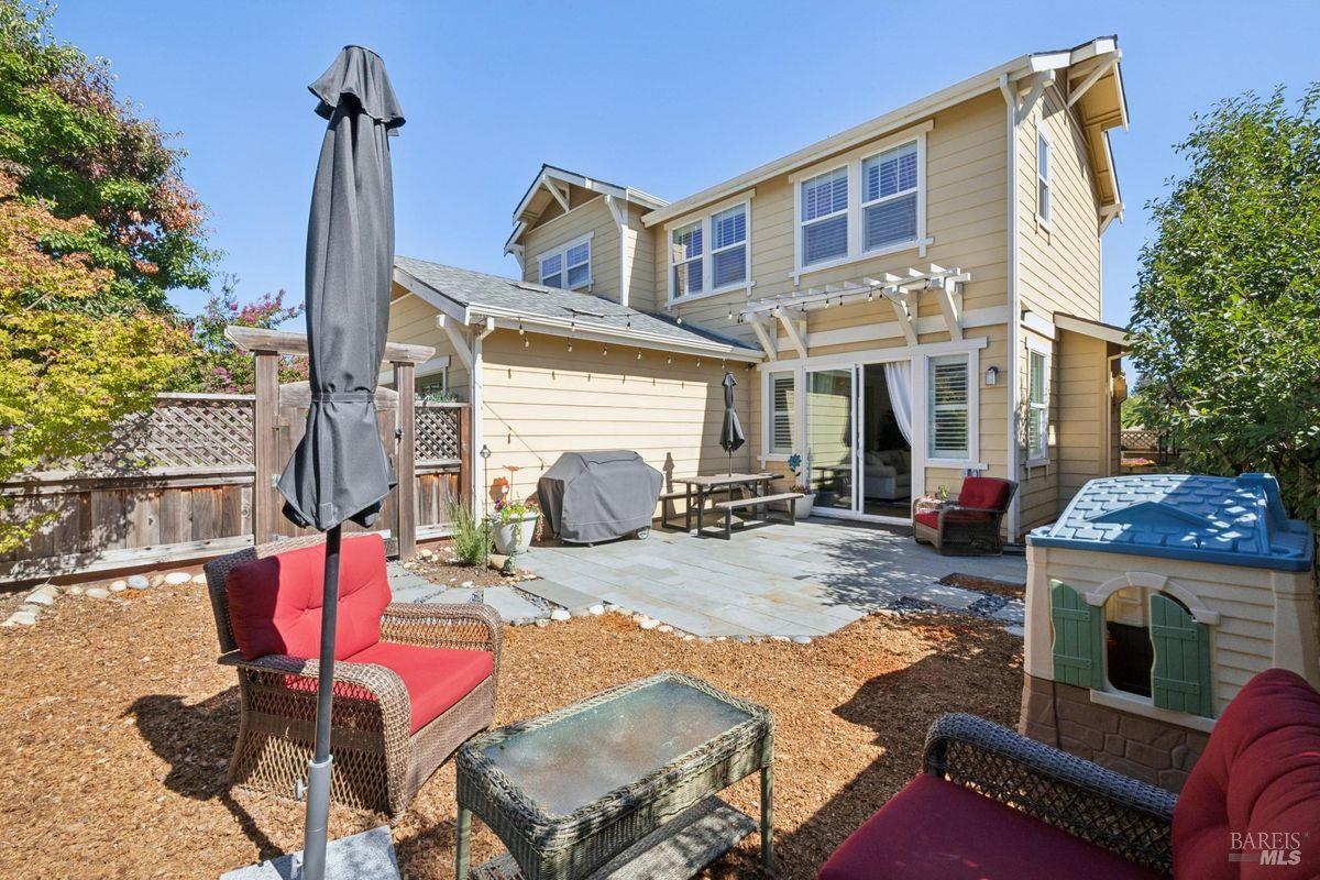 Two-story beige home viewed from the backyard, with a paved patio, outdoor seating, and fenced yard.