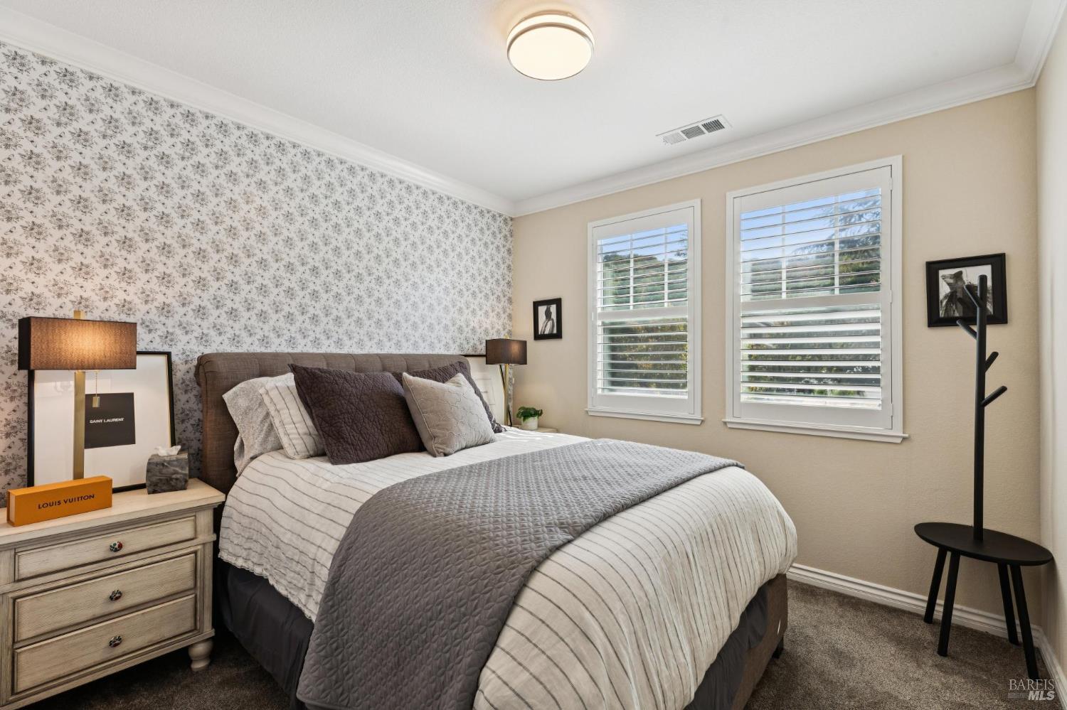 Bedroom with a large gray-quilted bed, floral wallpaper, and two shuttered windows.