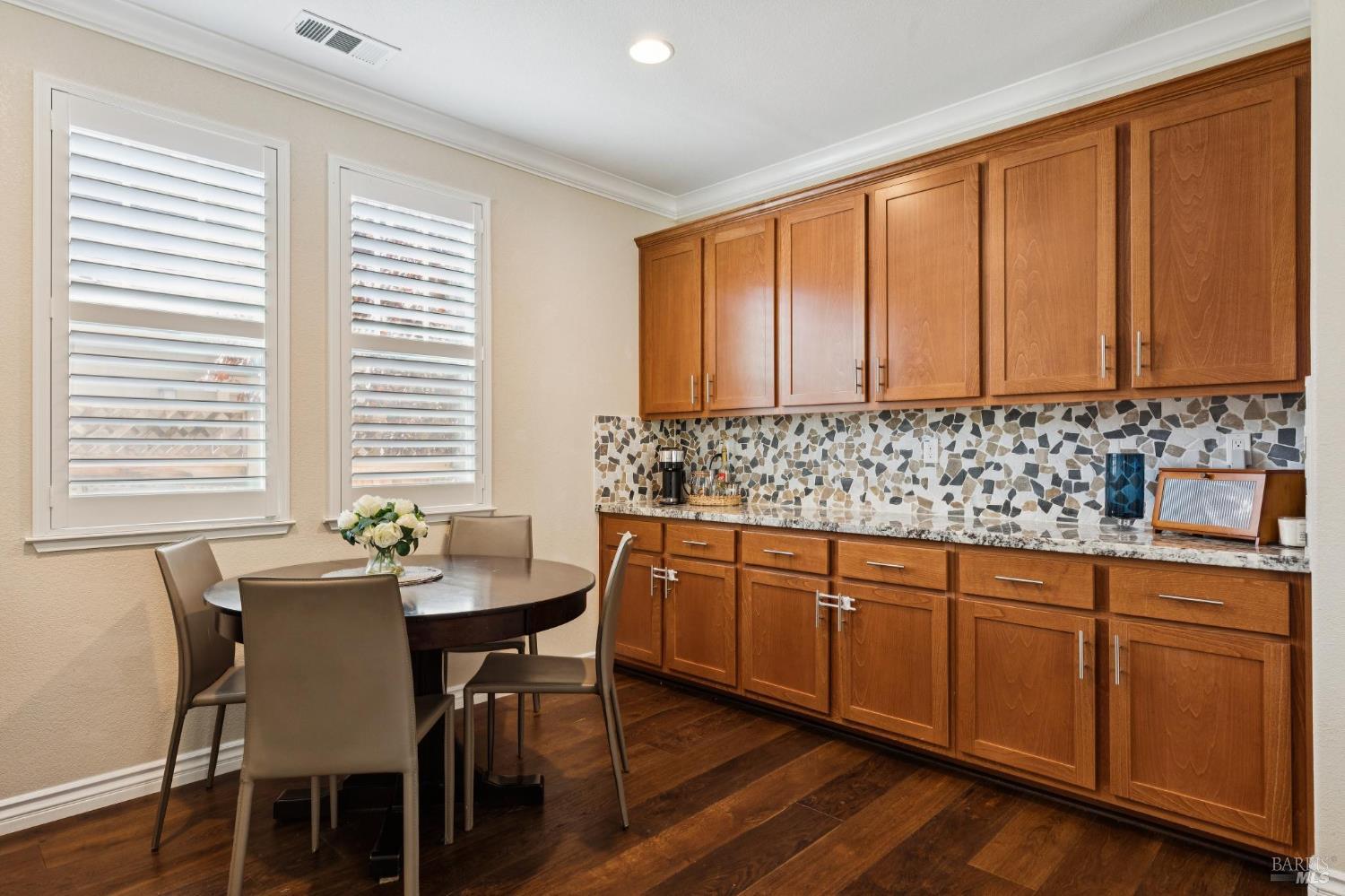 Kitchen and dining nook with wood cabinets, mosaic backsplash, and a round table by shuttered windows.