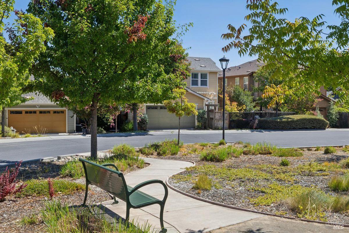 Street-facing homes behind a landscaped sidewalk, framed by mature trees and a green park bench.