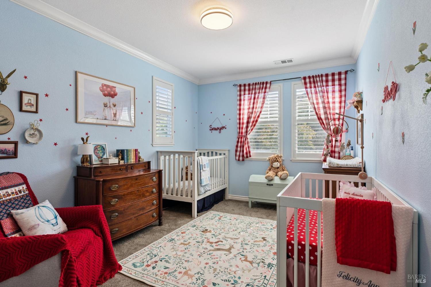 Nursery with two white cribs, red gingham curtains, and a wooden dresser.