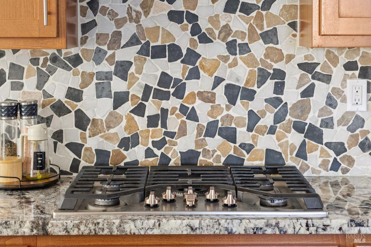 Kitchen gas range beneath an irregular stone backsplash, with wooden cabinets and a speckled stone countertop.