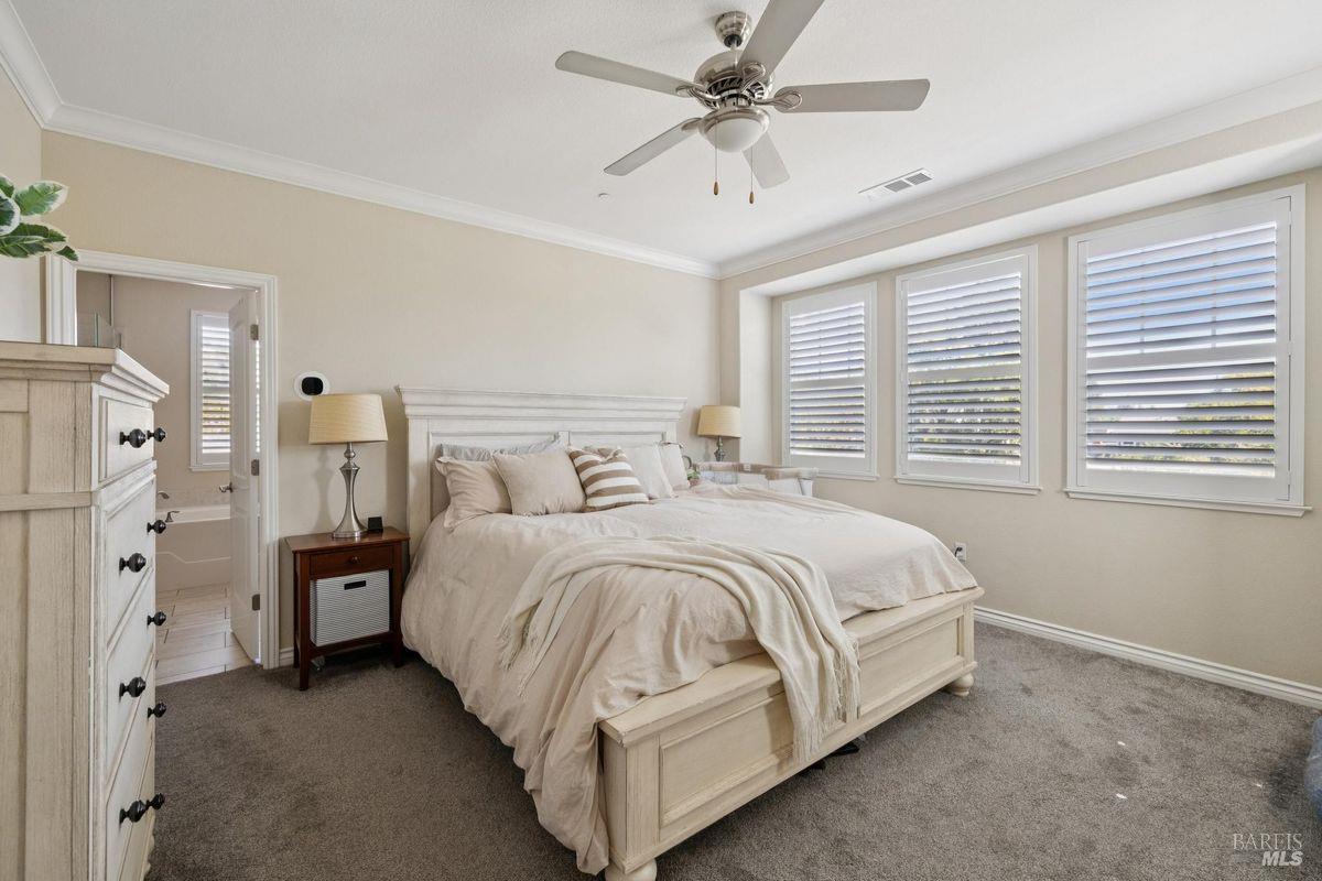 Bedroom with a large light-wood bed, ceiling fan, and three windows with white shutters.