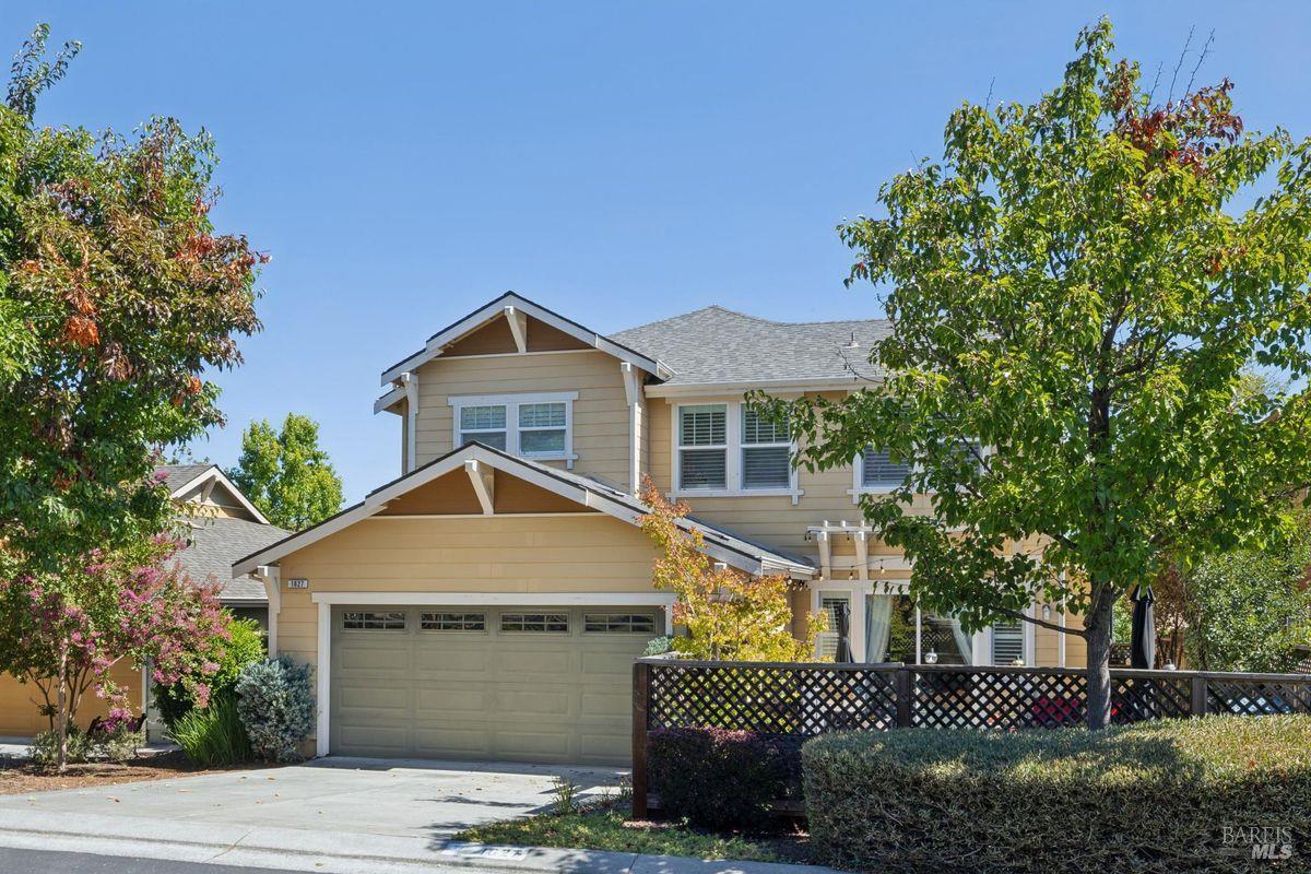 Two-story beige house with a two-car garage, lattice fence, and mature trees.