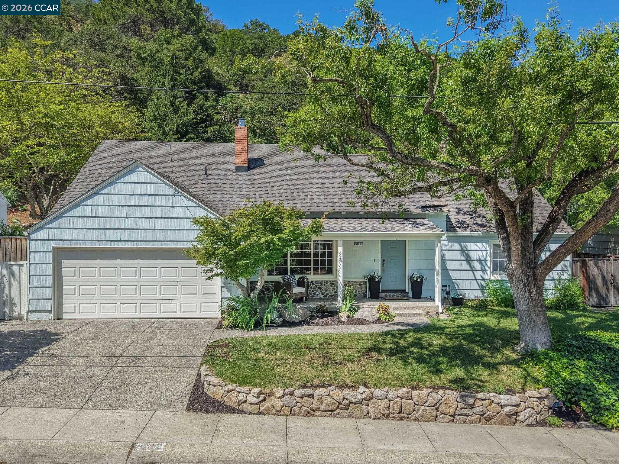 Blue-gray house with a two-car garage, covered entry, and mature trees shading the front yard.