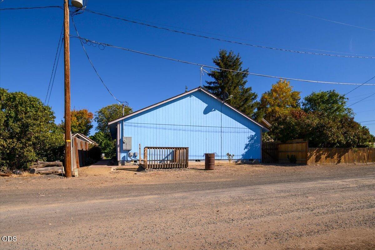 Blue gable-roof building beside a gravel road, with a small wooden deck and fenced yard.