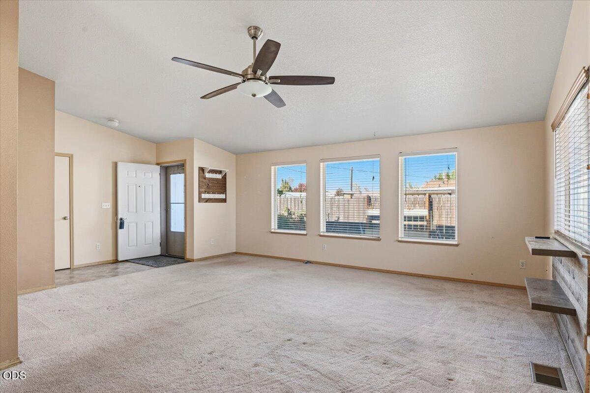 Empty living room with beige carpet, ceiling fan, and three windows overlooking a fenced yard.