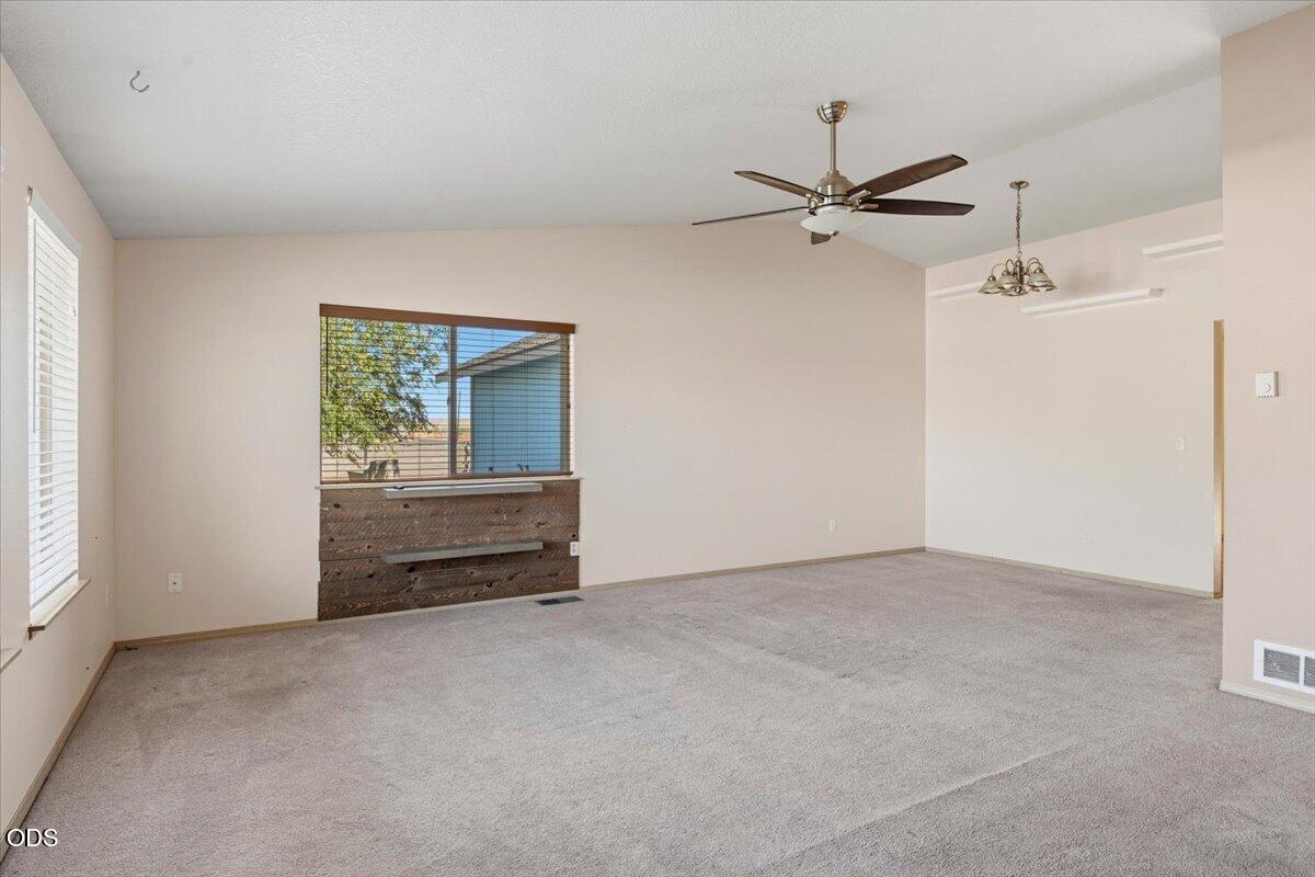 Vacant living room with gray carpet, a large window, dark wood wall panel, and ceiling fan.