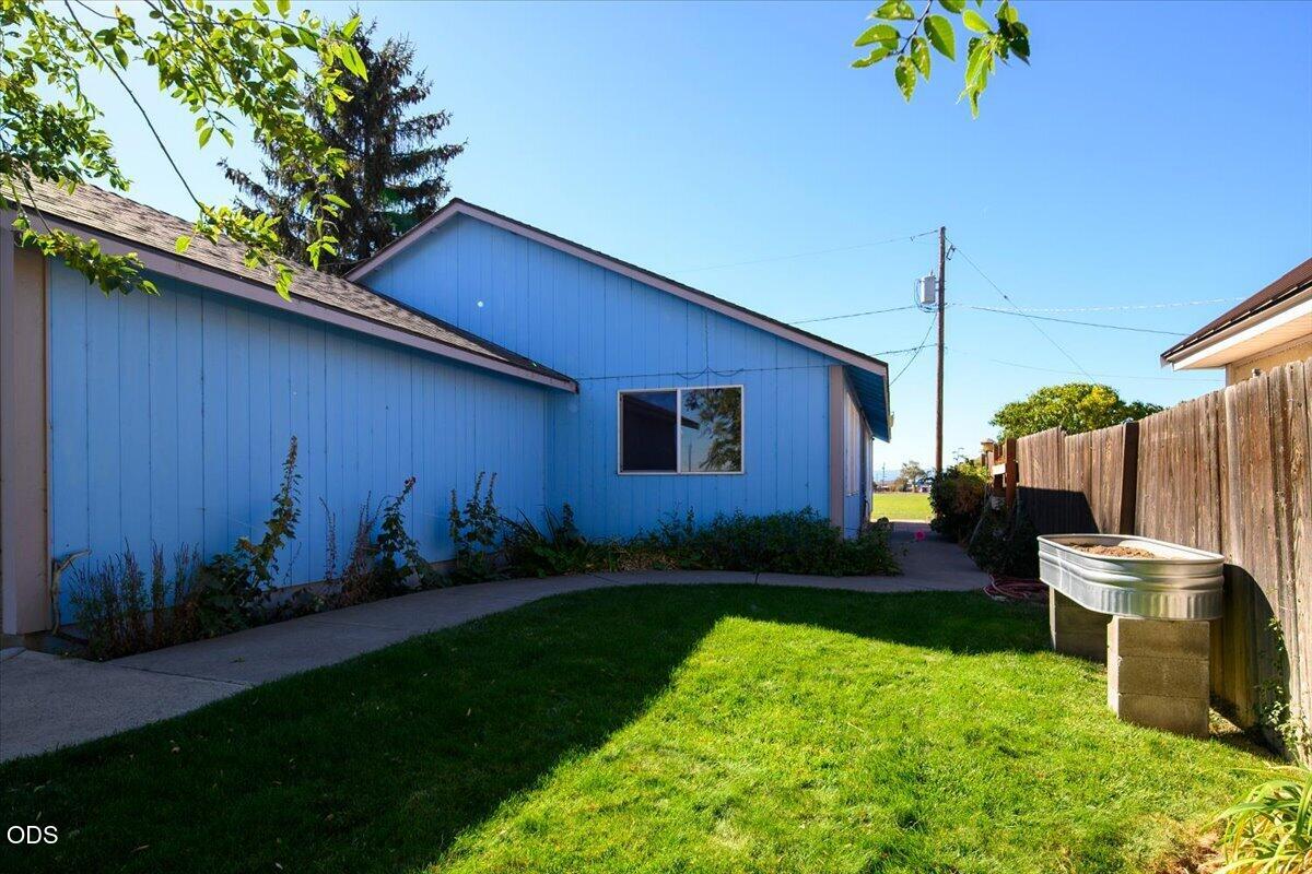 Blue-sided house beside a sunny lawn, concrete walkway, wooden fence, and metal trough planter.