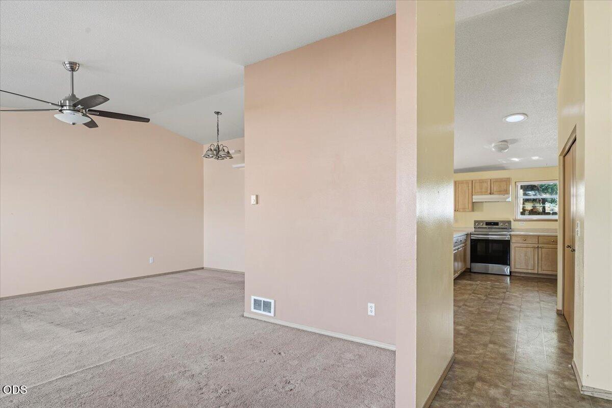 Empty carpeted living room opening into a tiled kitchen with wood cabinets and stainless steel appliances.