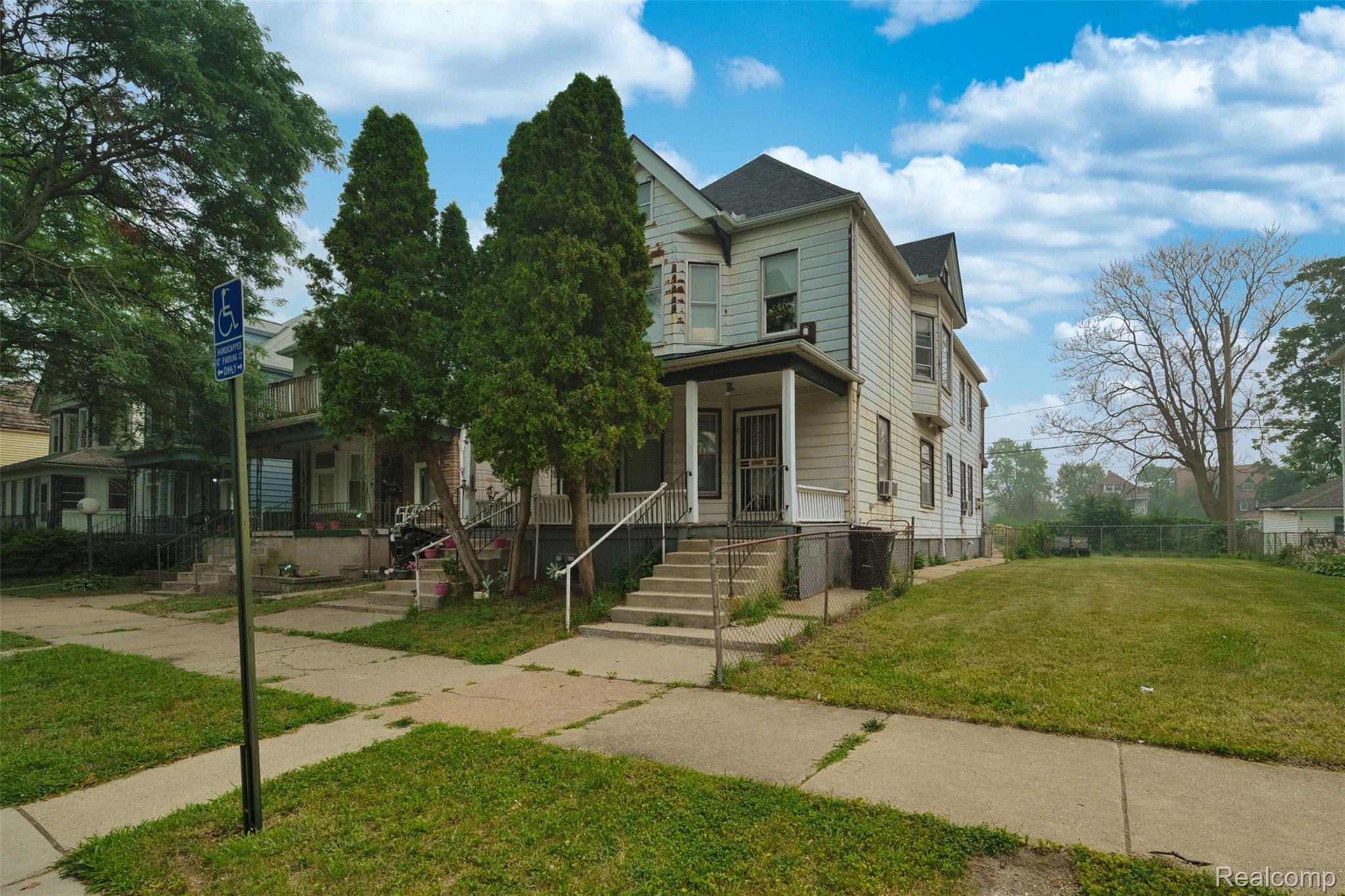 Two-story light-sided house with a covered porch, front steps, and mature evergreen trees.