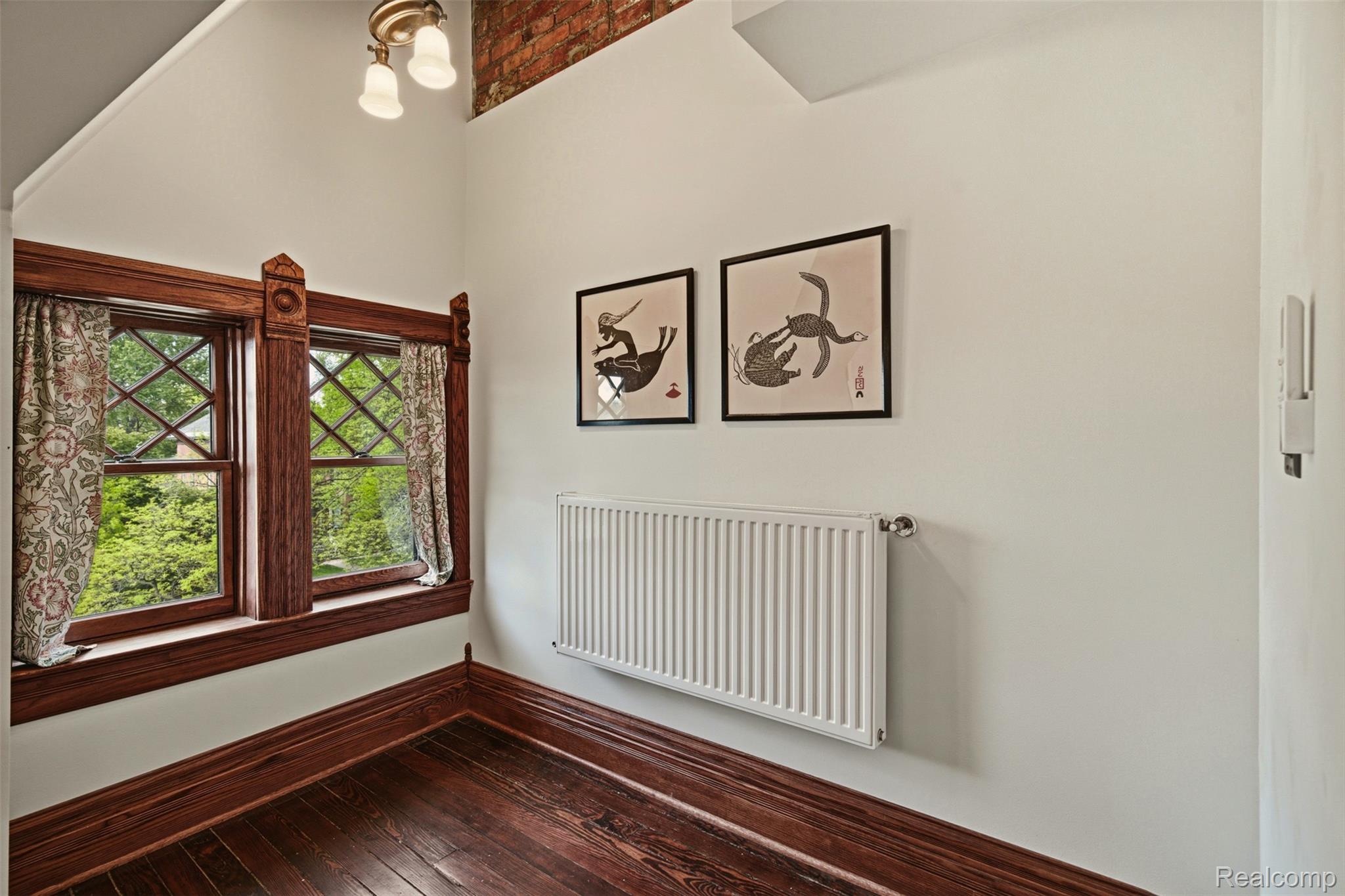 Room with dark hardwood floors, ornate wood-trimmed windows, patterned curtains, and a wall radiator.