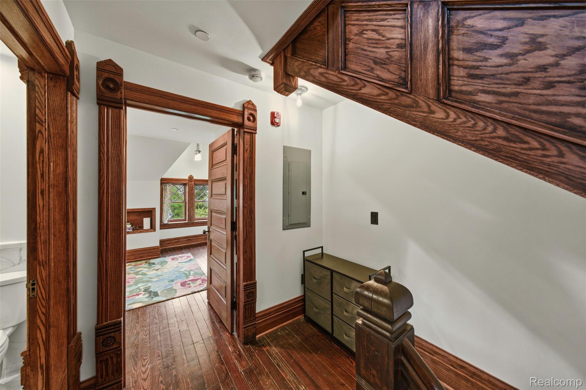 Hallway with ornate dark wood trim, hardwood floors, and an open door to a room with a floral rug.