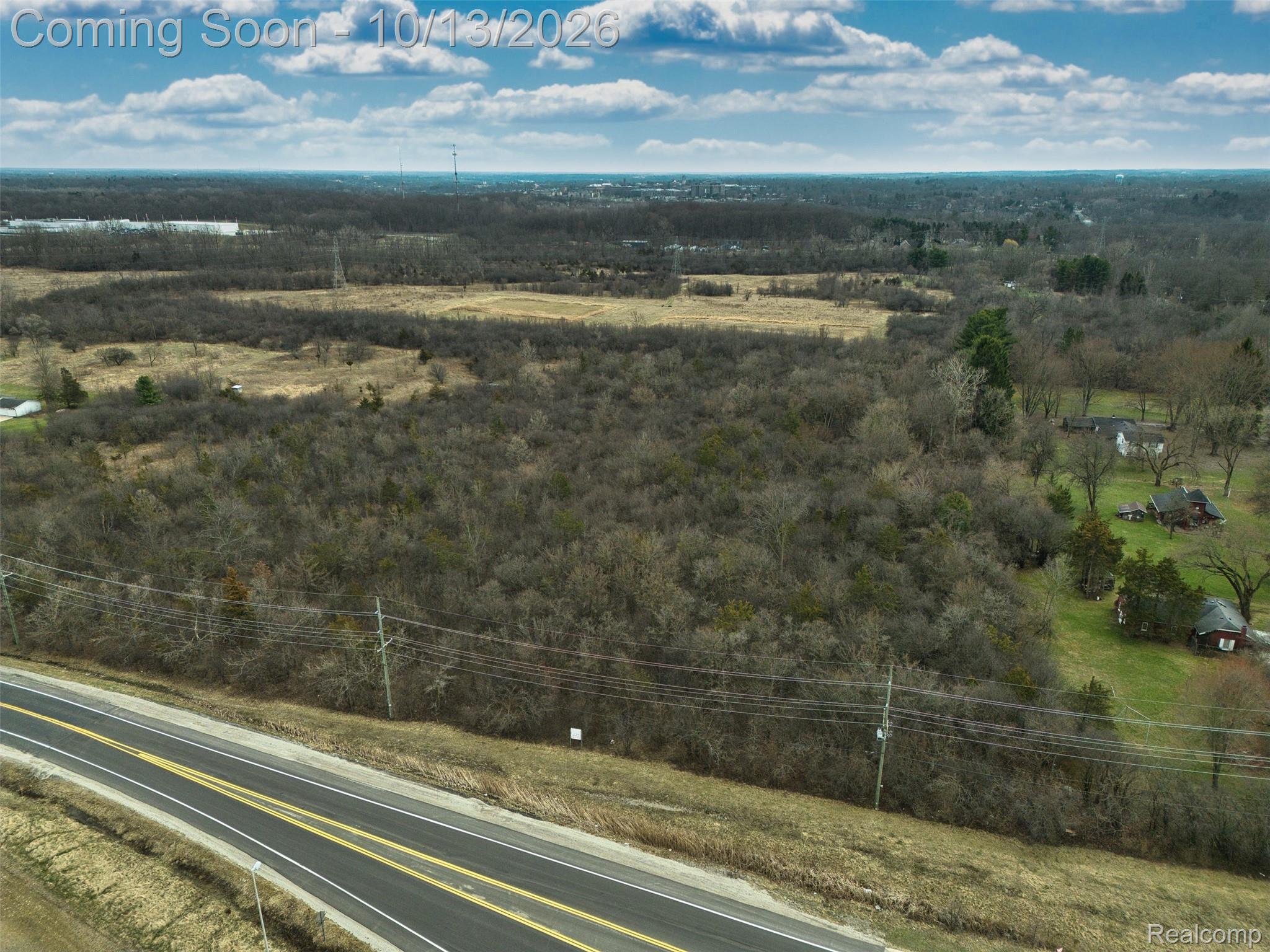 Aerial view of a wooded vacant parcel beside a two-lane road, with fields and homes nearby.