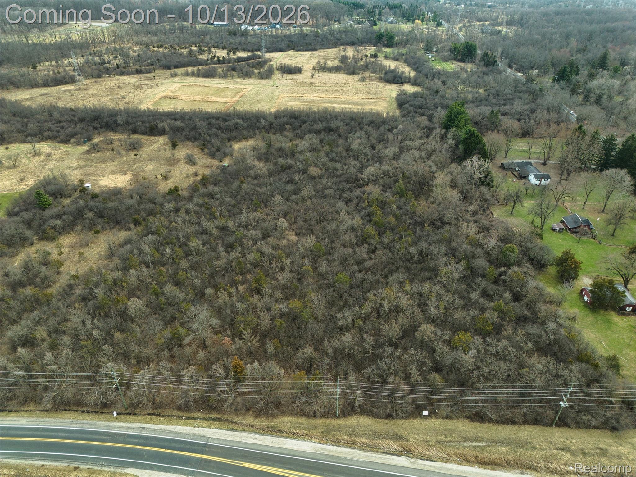 Aerial view of wooded land with open fields, nearby homes, and road frontage.