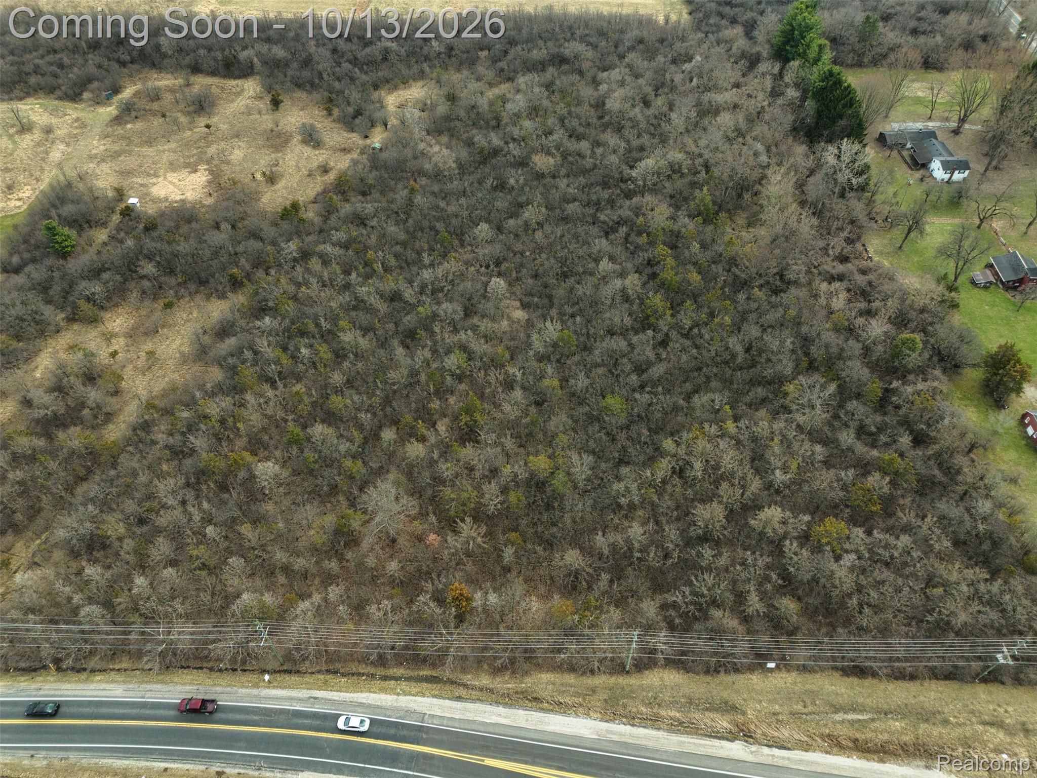 Aerial view of a wooded vacant parcel along a paved road, with utility lines and neighboring homes.