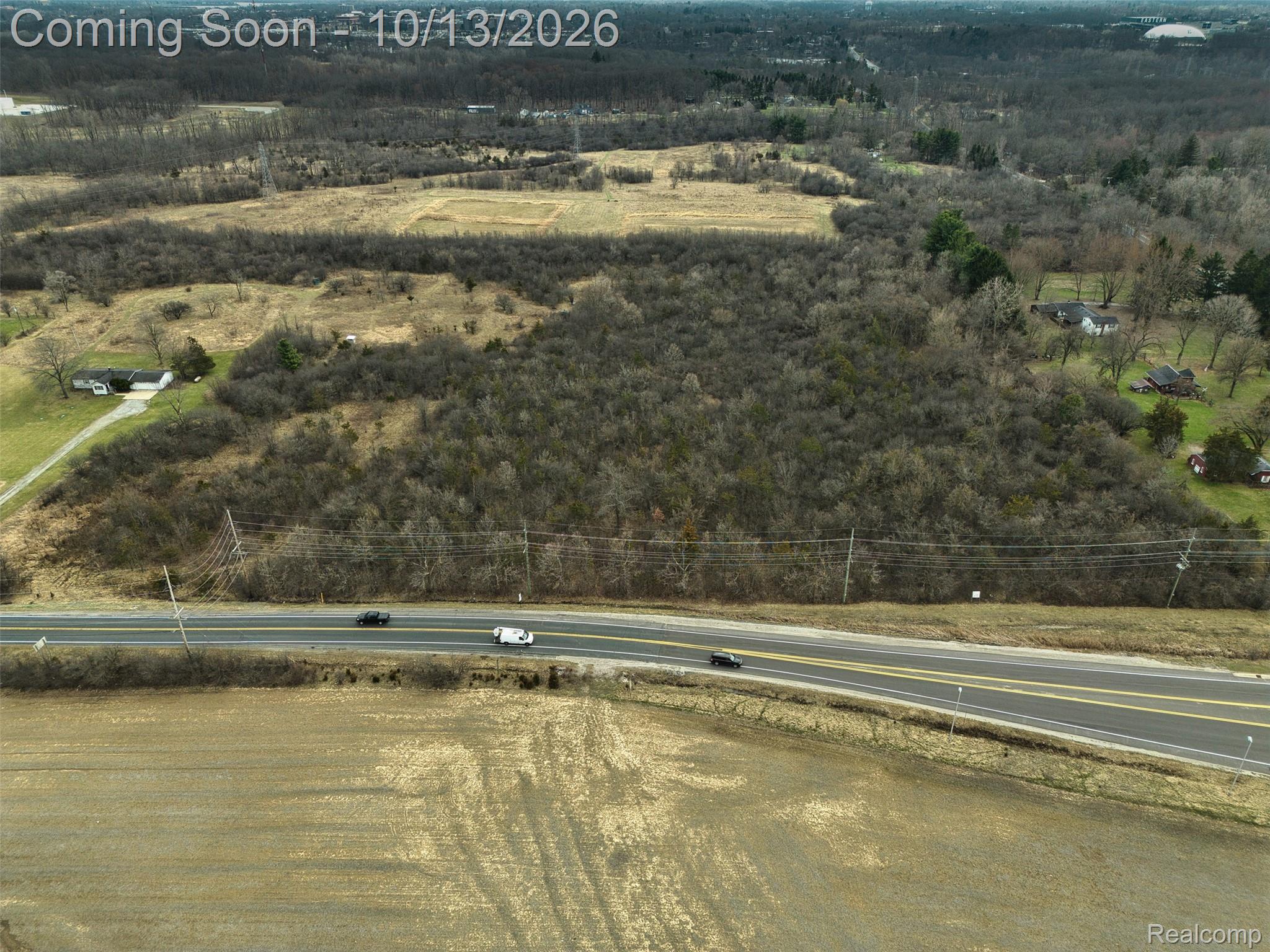 Aerial view of wooded vacant land and open fields beside a paved road.