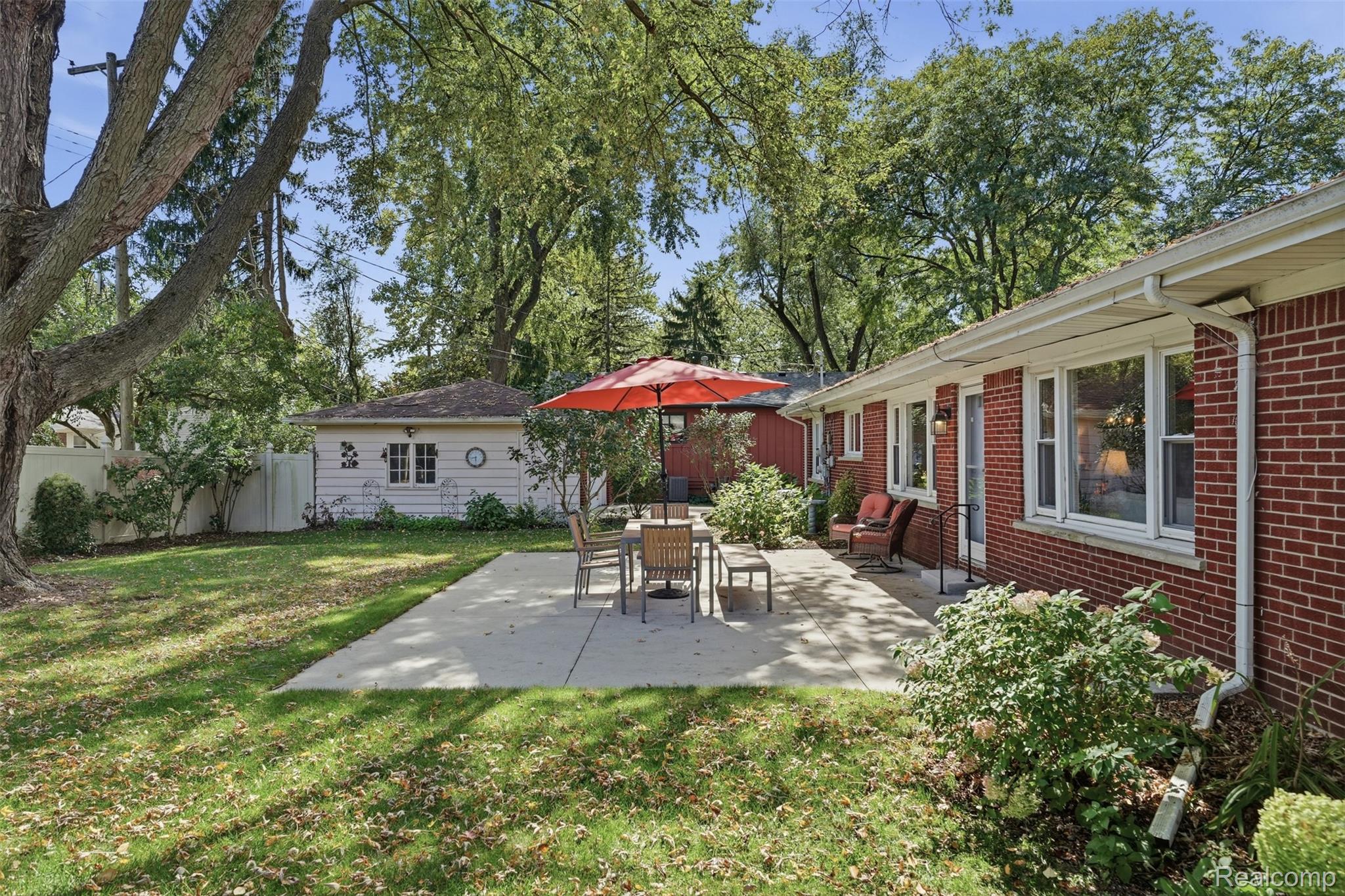 Backyard patio beside a red brick house, furnished with outdoor dining chairs beneath a red umbrella.
