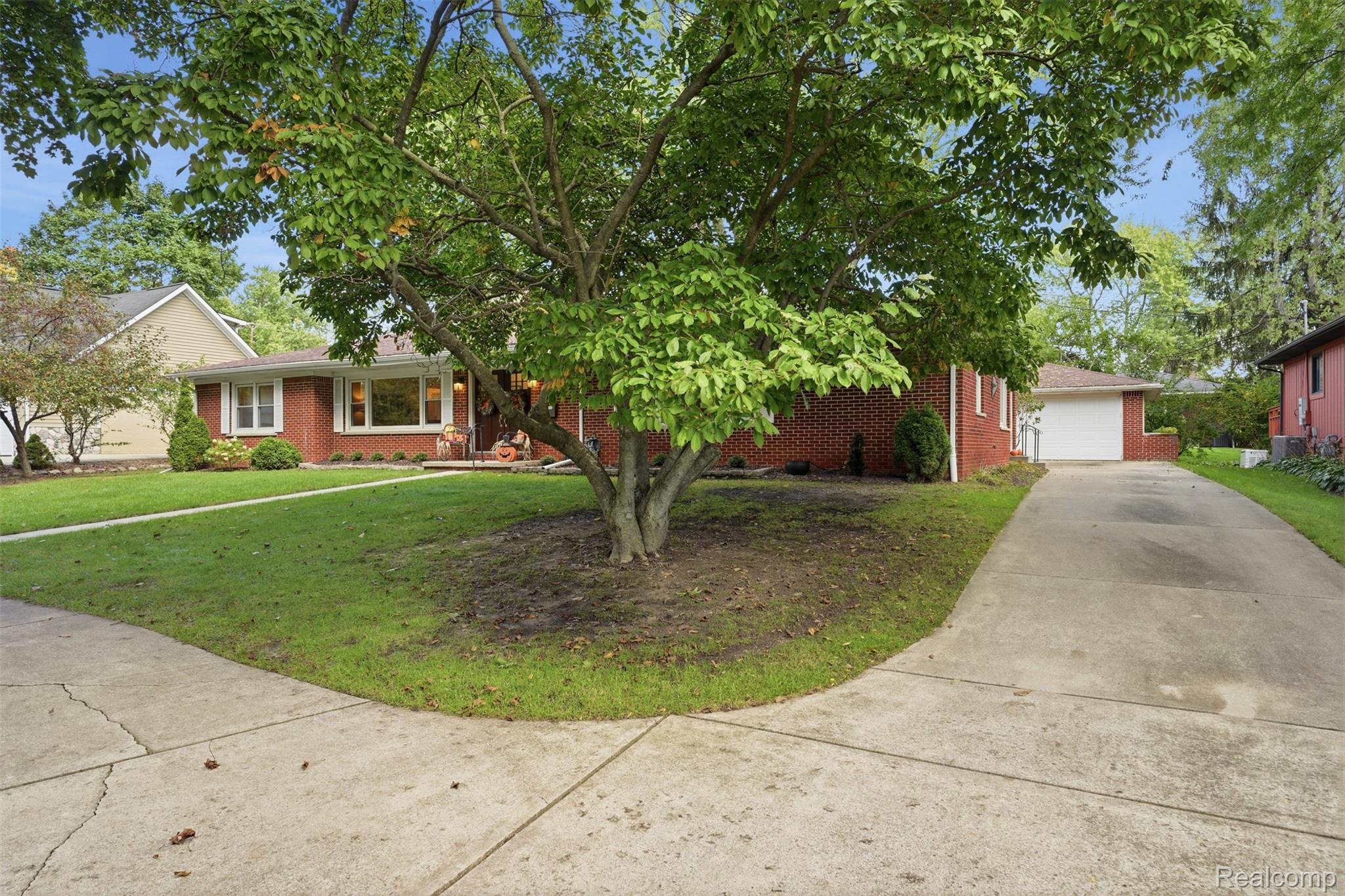 Red-brick ranch home partly shaded by a large tree, with a wide driveway leading to the attached garage.