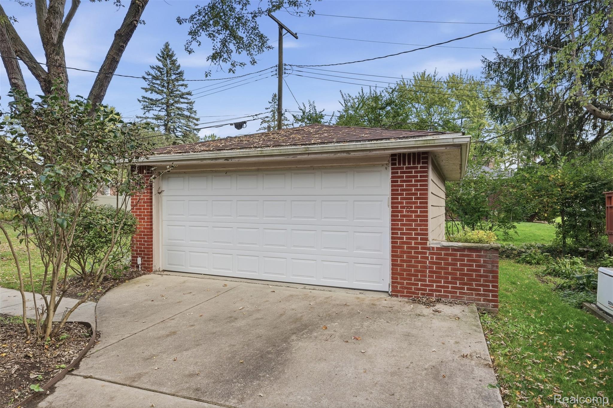 Red-brick garage with a wide white double door and concrete driveway amid leafy landscaping.