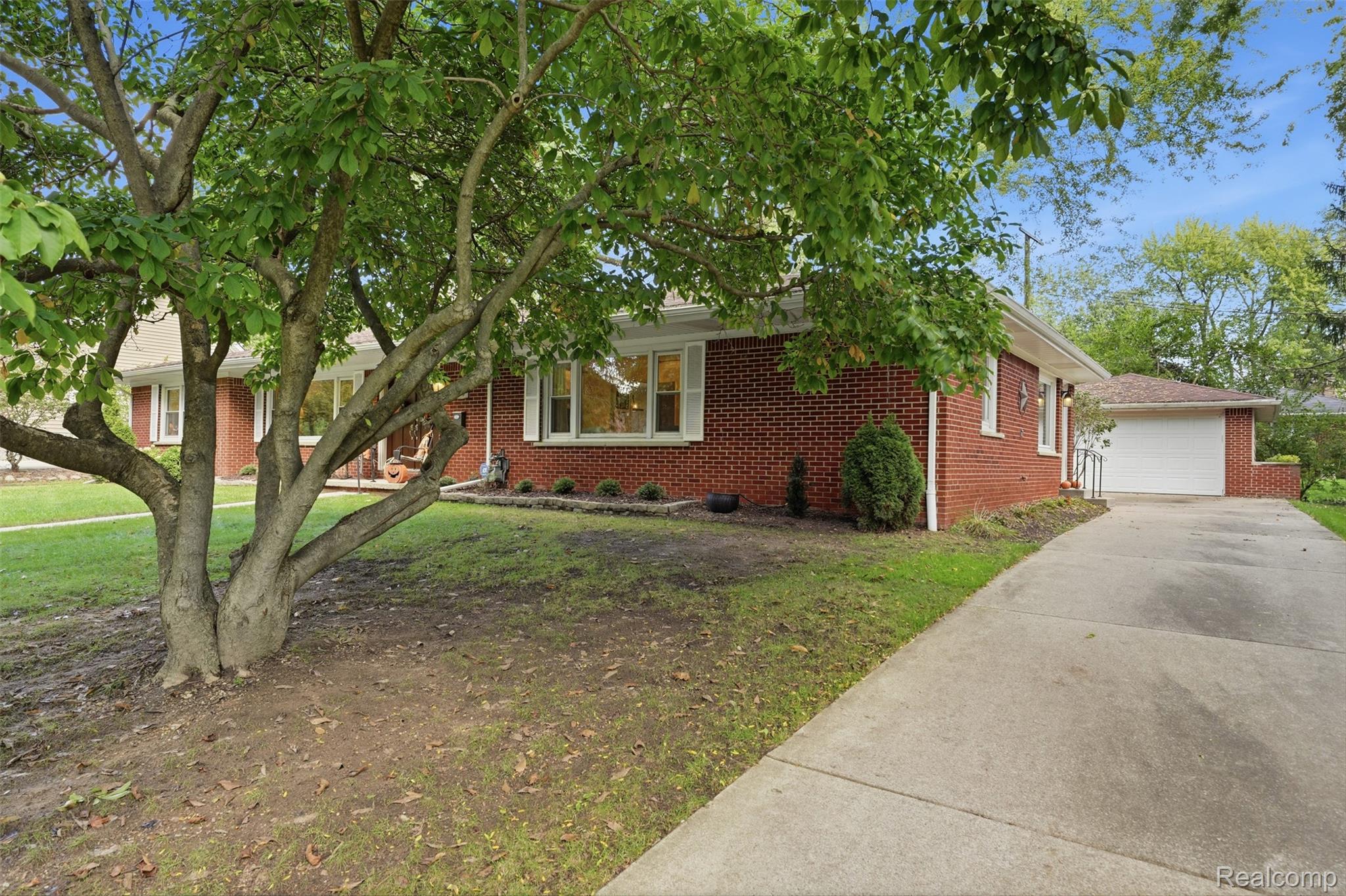 Red brick ranch house and garage behind a large leafy tree, with a concrete driveway.