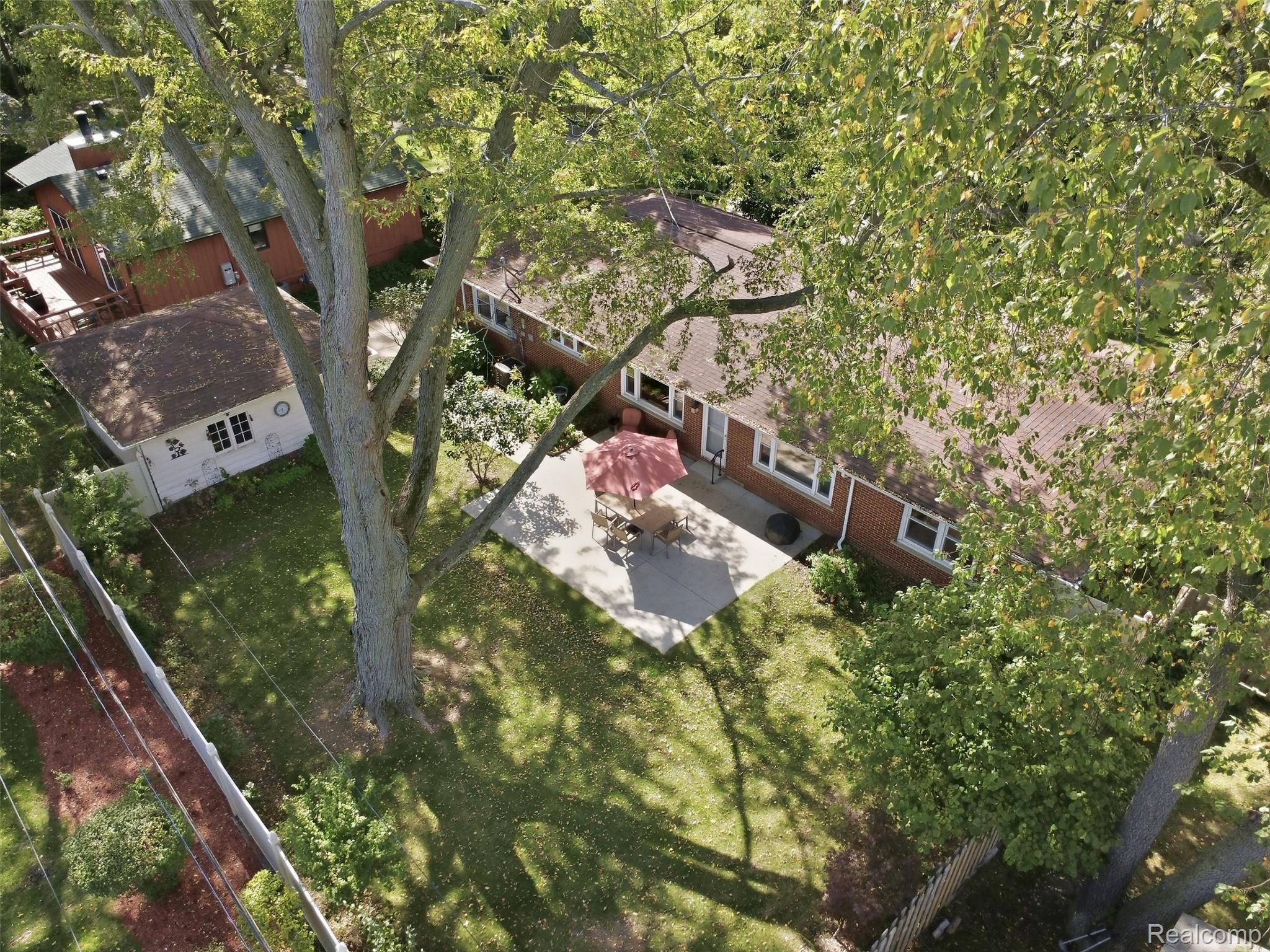 Aerial view of a brick house, leafy yard, and patio with outdoor dining furniture and a red umbrella.