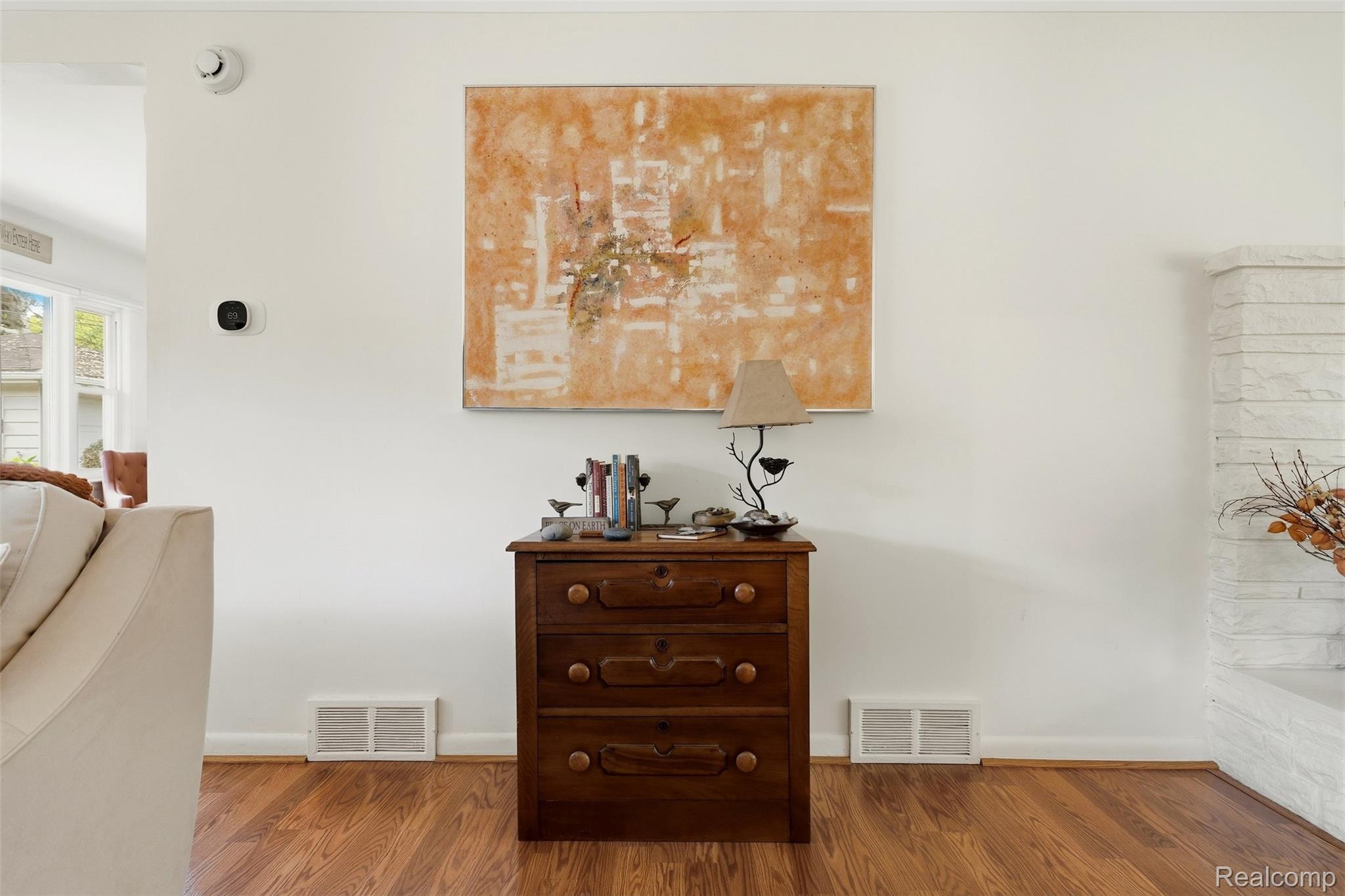 Living room with a wooden chest beneath orange abstract art, hardwood floors, and a white stone fireplace.