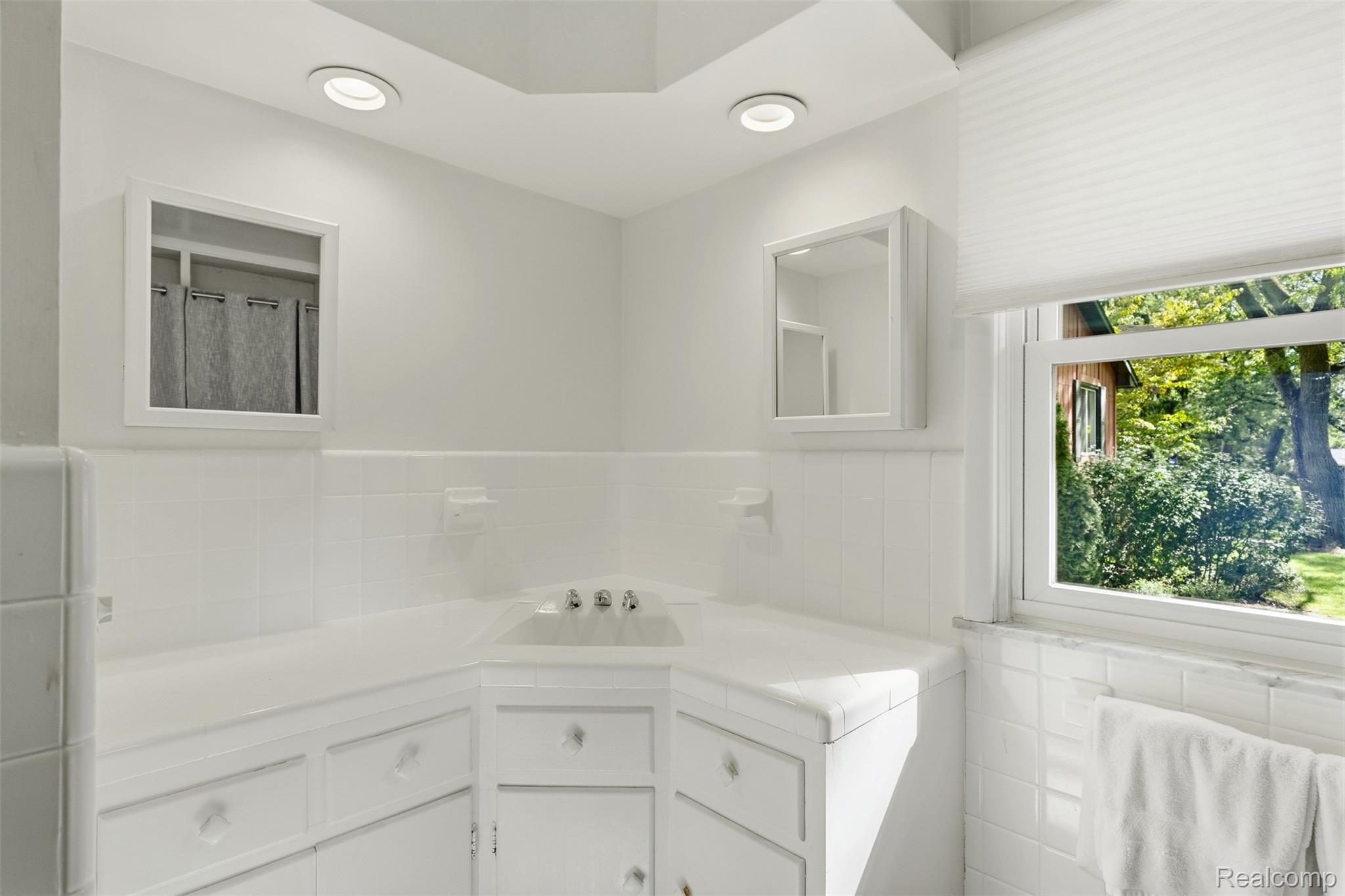 White tiled bathroom vanity with two mirrors and a window overlooking leafy greenery.