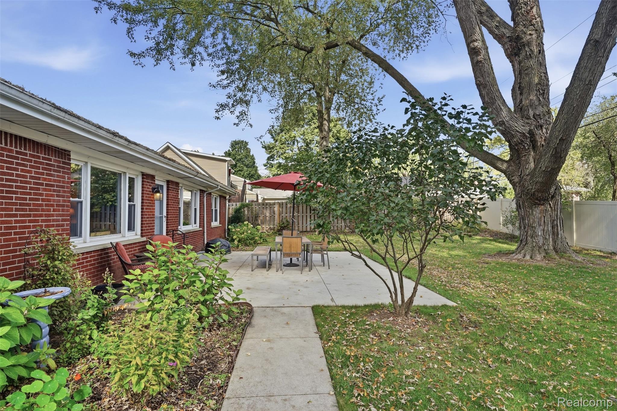 Backyard patio with outdoor dining set, red umbrella, garden beds, and a large shade tree.