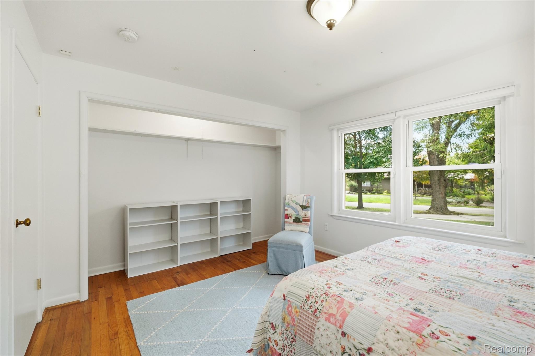 Bedroom with a floral patchwork bedspread, hardwood floors, and large windows overlooking leafy trees.
