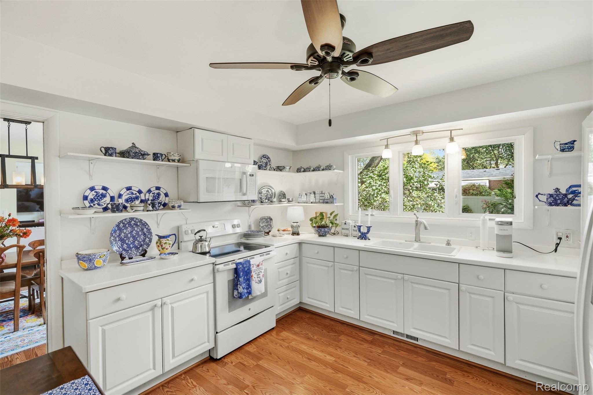 Bright white kitchen with blue-and-white dishes on open shelves and a sink beneath wide windows.