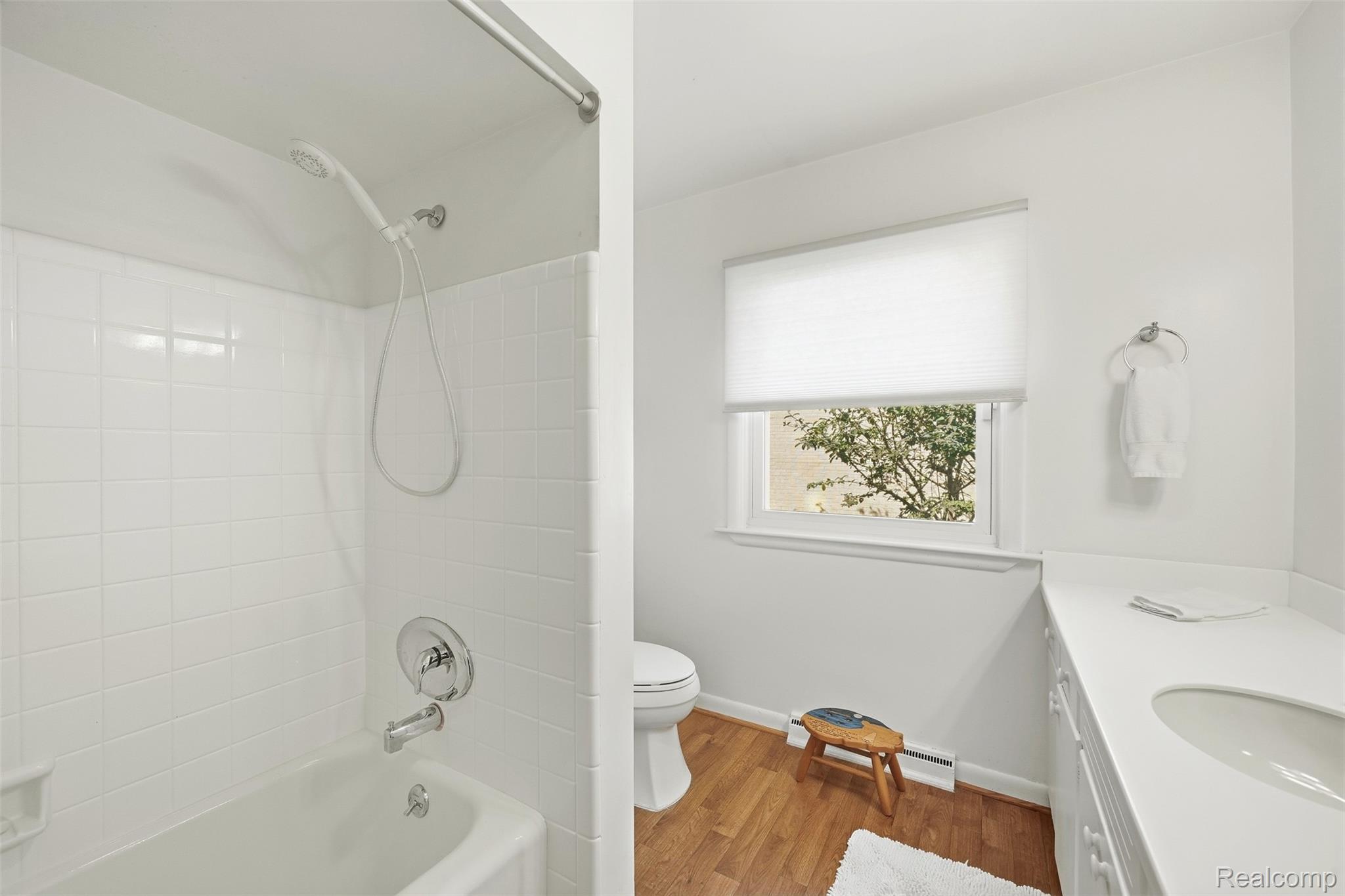 Bathroom with white tiled tub and shower, wood flooring, window, toilet, and vanity.