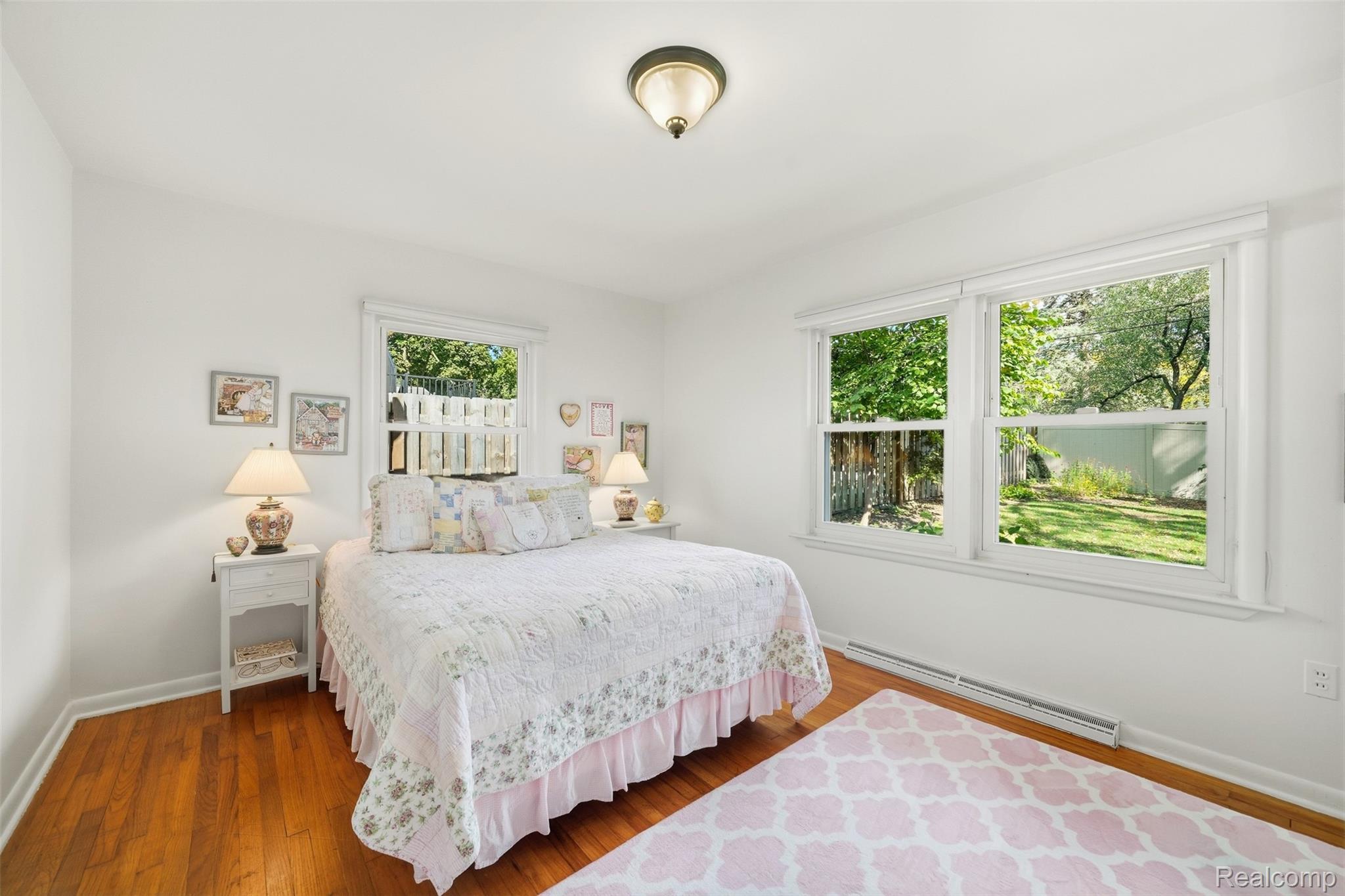 Bedroom with a white patchwork bedspread, hardwood floors, and large windows overlooking a leafy yard.