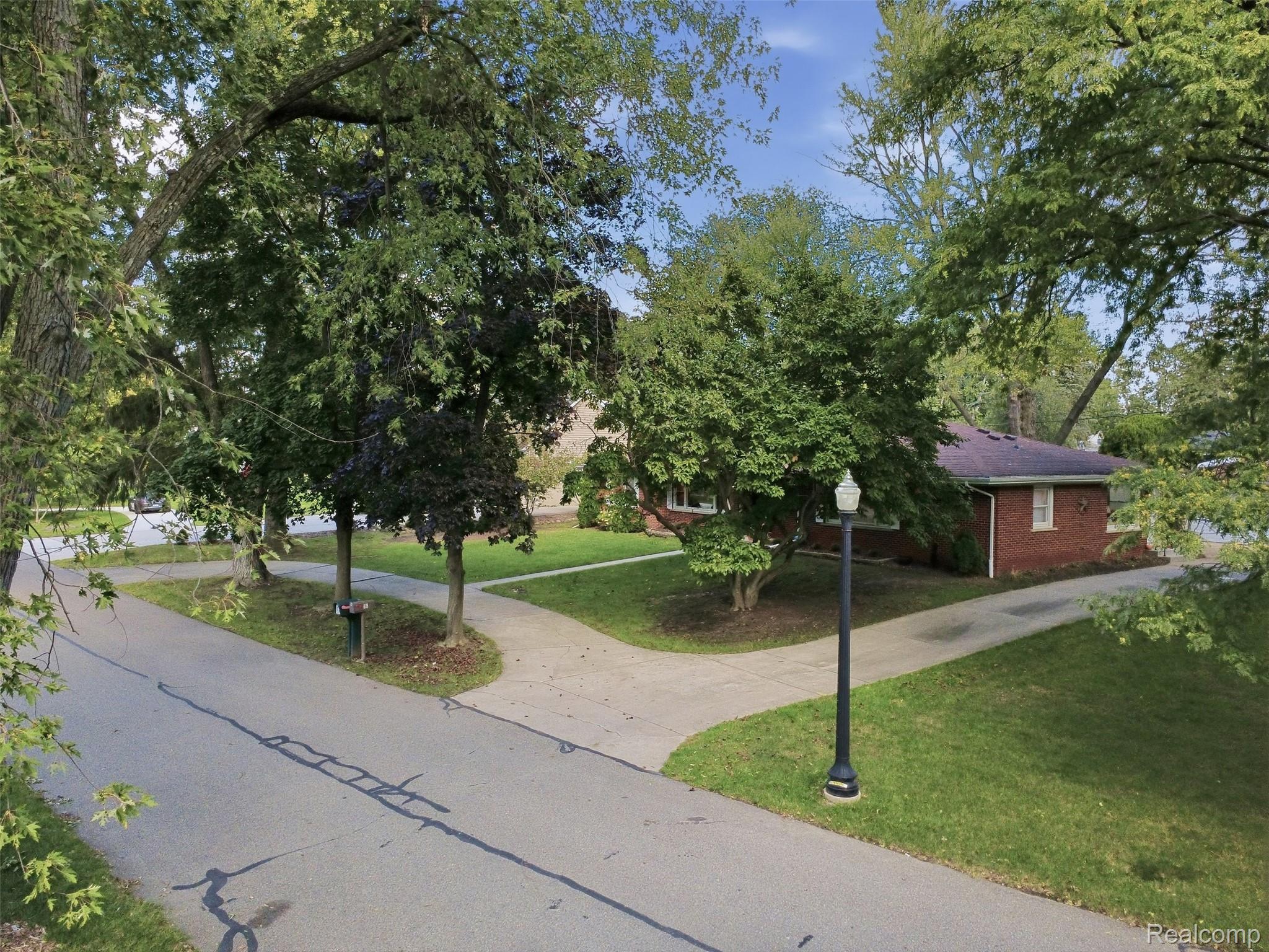 Red-brick house partly screened by mature trees, with a corner lawn, sidewalk, and lamppost.